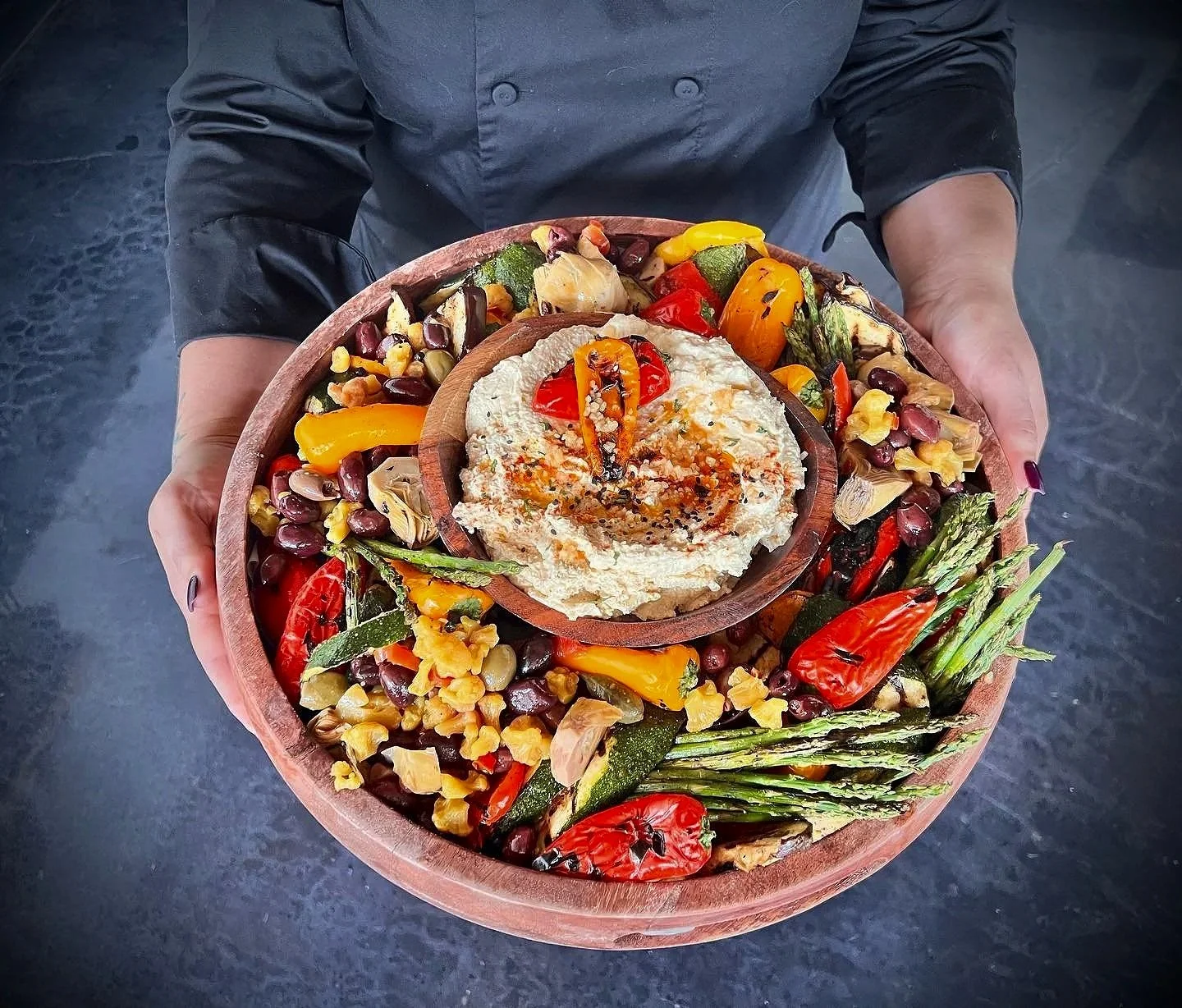 A person holding a large wooden bowl filled with assorted roasted vegetables, including red and yellow peppers, zucchini, asparagus, and cherry tomatoes. A smaller wooden bowl with hummus is placed on top of the larger bowl, garnished with a cherry tomato, red pepper, and olive oil.