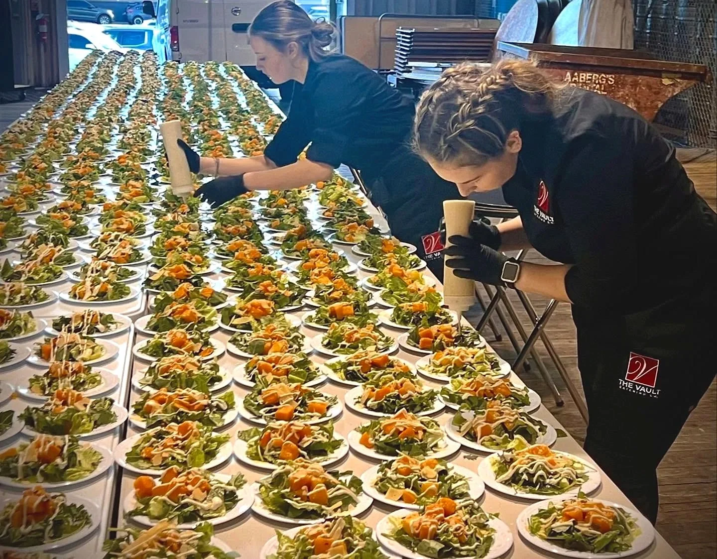 Two women wearing black uniforms and gloves preparing numerous salads on plates at a long table in a restaurant or event space.