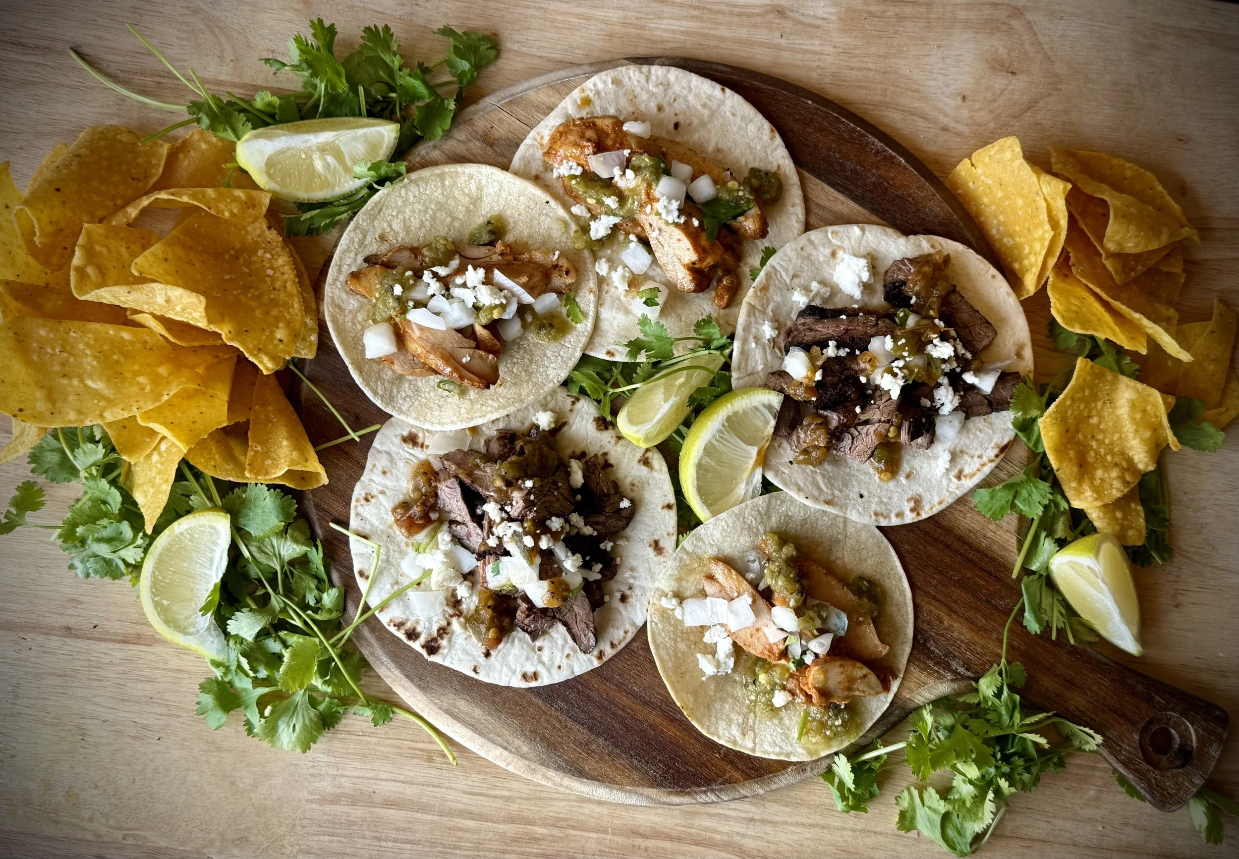 A wooden platter with five tacos topped with various meats, chopped onions, and cheese, garnished with lime wedges, cilantro, and tortilla chips on a light wooden table.
