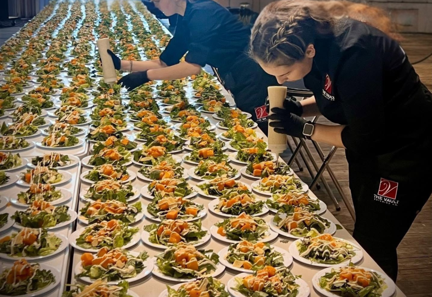 Two people preparing numerous plates of salad on a long table. They are wearing black gloves and black uniforms with a logo that says 'THE VAULT.' The plates are filled with leafy greens, cubed yellow vegetables, and drizzled dressing.