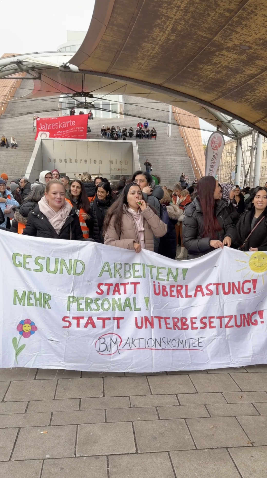 Menschenkette mit einer Protestaktion, bei der eine Frau eine Botschaft auf einem Banner trägt, das für bessere Arbeitsbedingungen und gegen Überlastung und Unterbesetzung protestiert. Die Menschenmenge steht vor einer Treppe und mehreren Stufen mit Zuschauern.