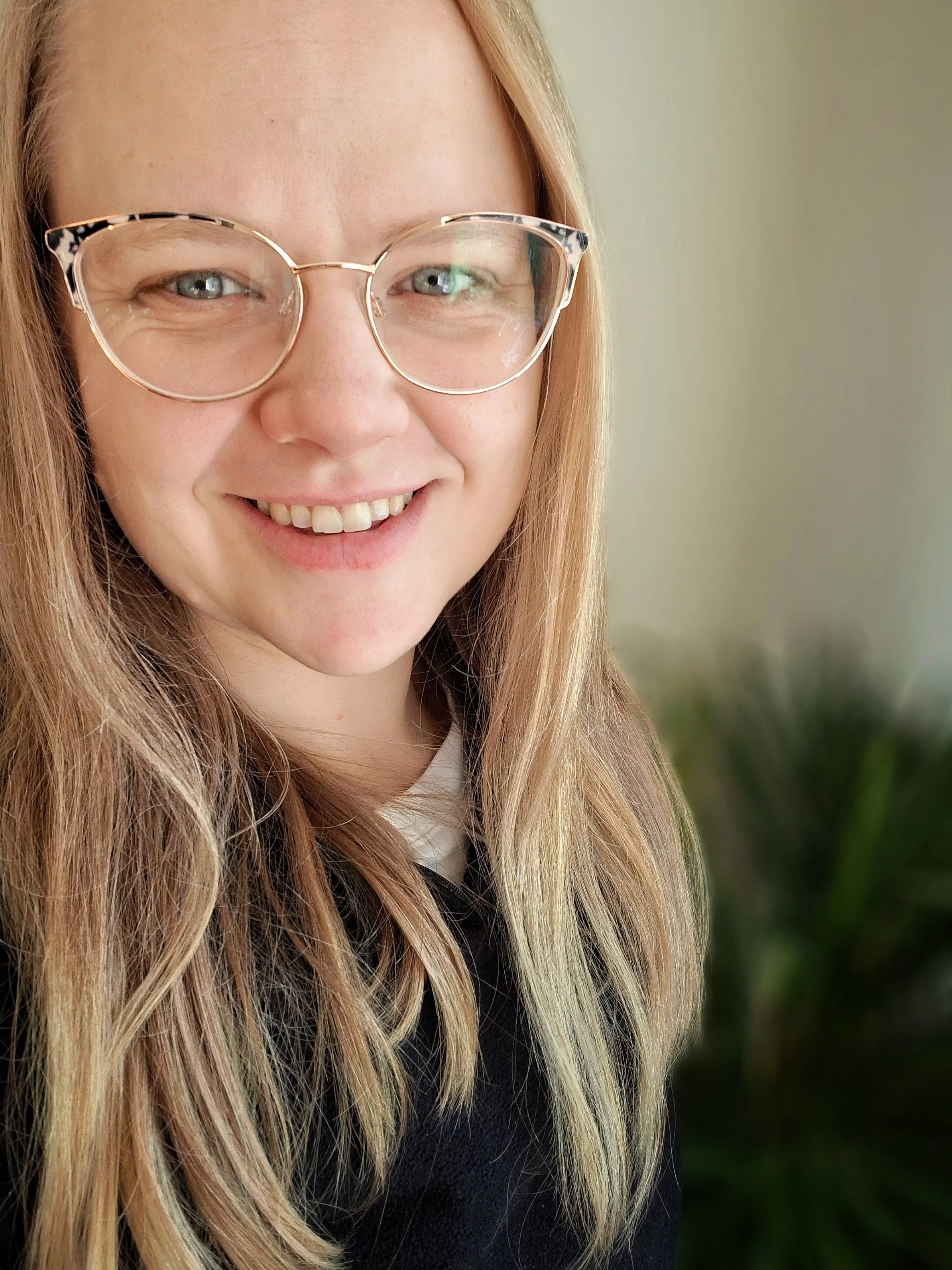 Close-up of a smiling woman with blonde hair, glasses, and light skin, wearing a black top and a white shirt, with a blurred green plant in the background. Doula and Antenatal Educator. hypnobirthing teacher.