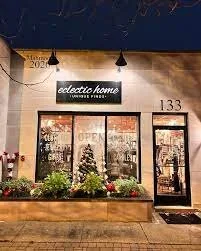 Exterior view of a store named 'eclectic home' with large windows and a door, decorated with plants and flowers in front, with string lights overhead.