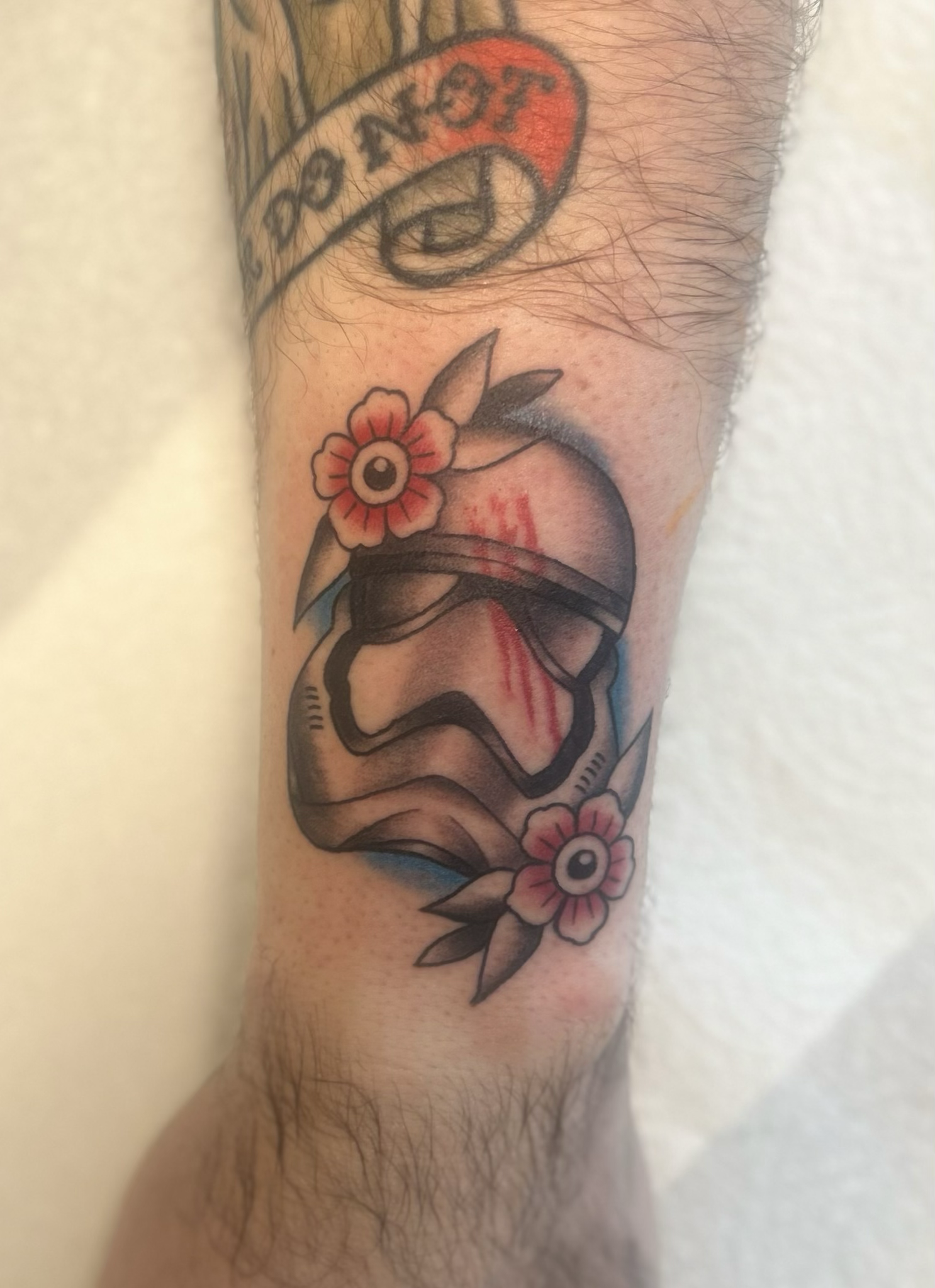 Neo‑traditional Stormtrooper helmet tattoo with red flowers, blue pop, bloody fingerprint detail