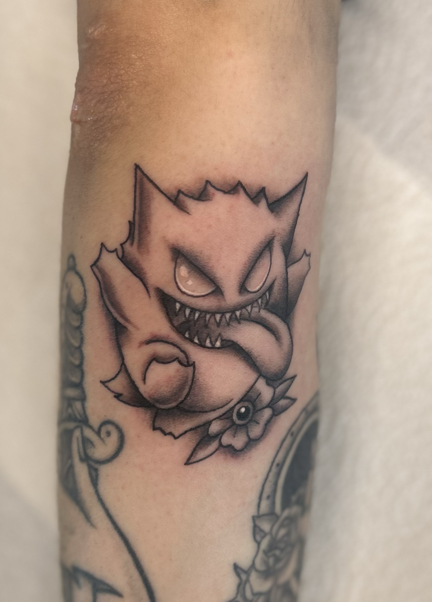 Black & gray custom Gengar Pokémon tattoo with sharp teeth and traditional flower accent