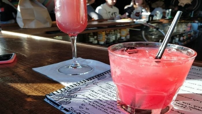 Two pink cocktails, one in a tall glass and the other in a short round glass with a straw, on a wooden bar counter with a group of people in the background.