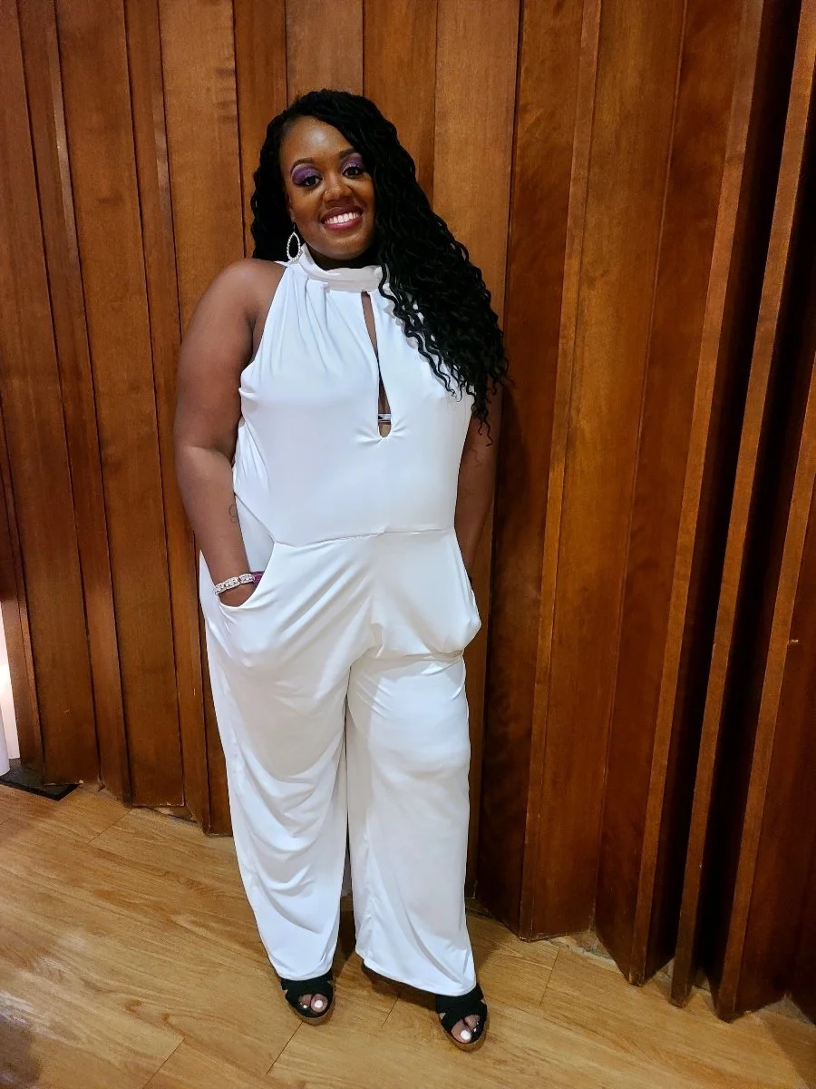 Woman wearing a white sleeveless jumpsuit with a keyhole neckline and black open-toe heels, standing against a wooden paneled wall, smiling.
