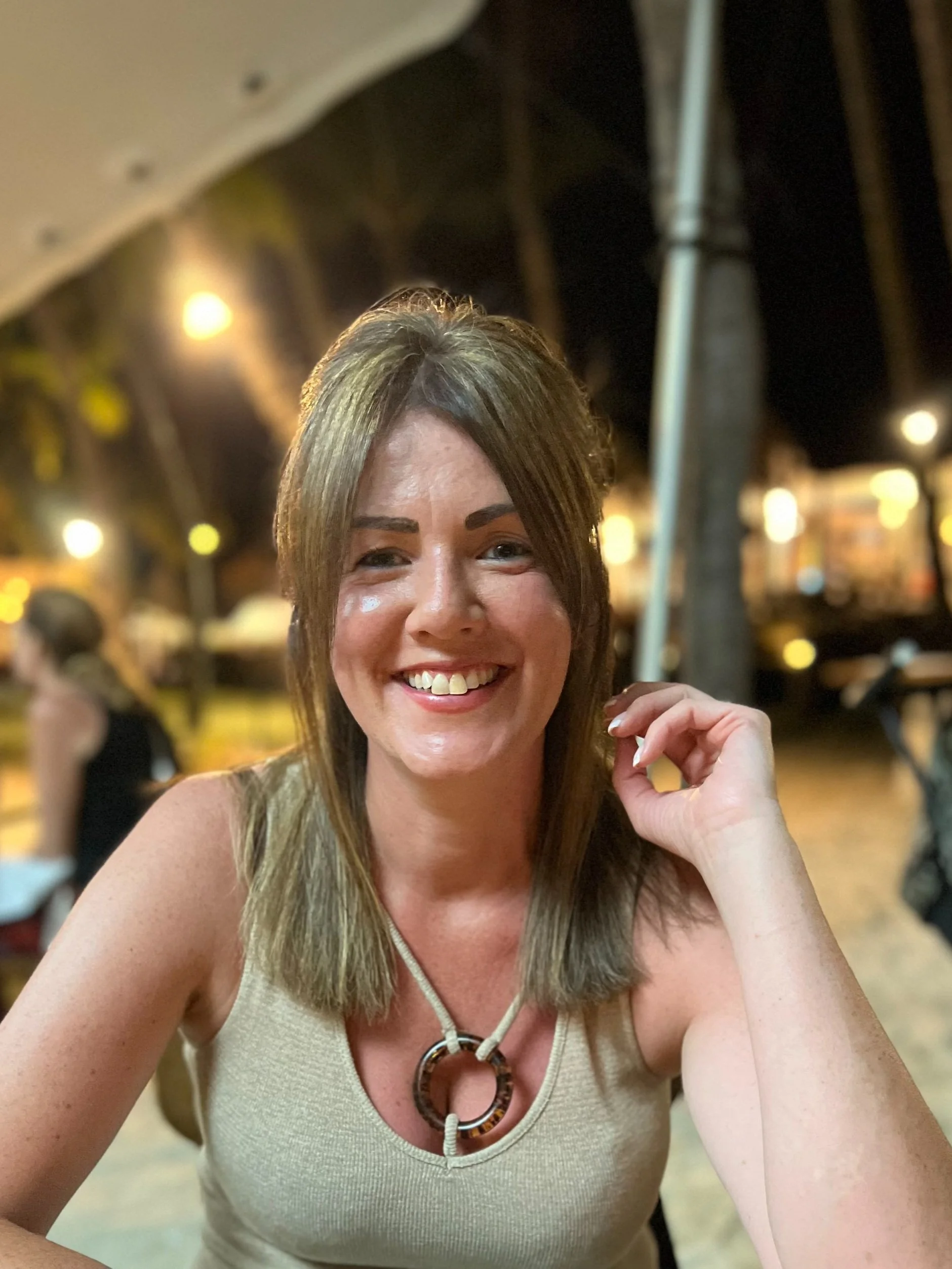 A woman with shoulder-length brown hair smiling at the camera at an outdoor restaurant or bar in the evening.
