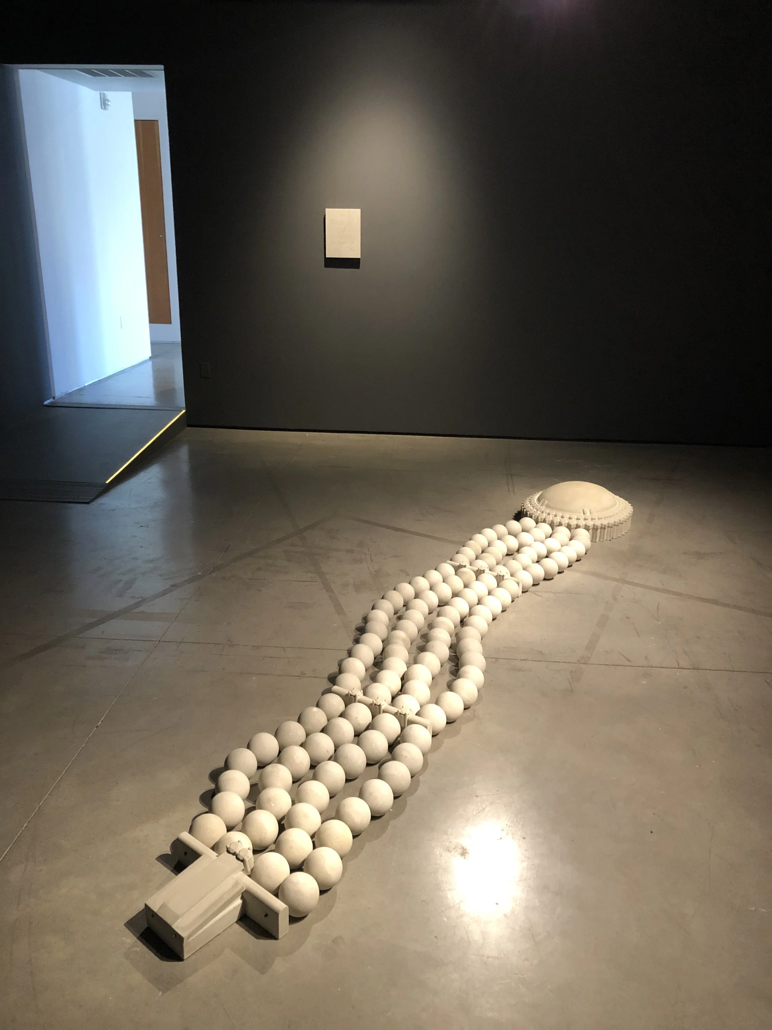 Contemporary art installation with rows of white spheres on the floor in a dimly lit gallery.