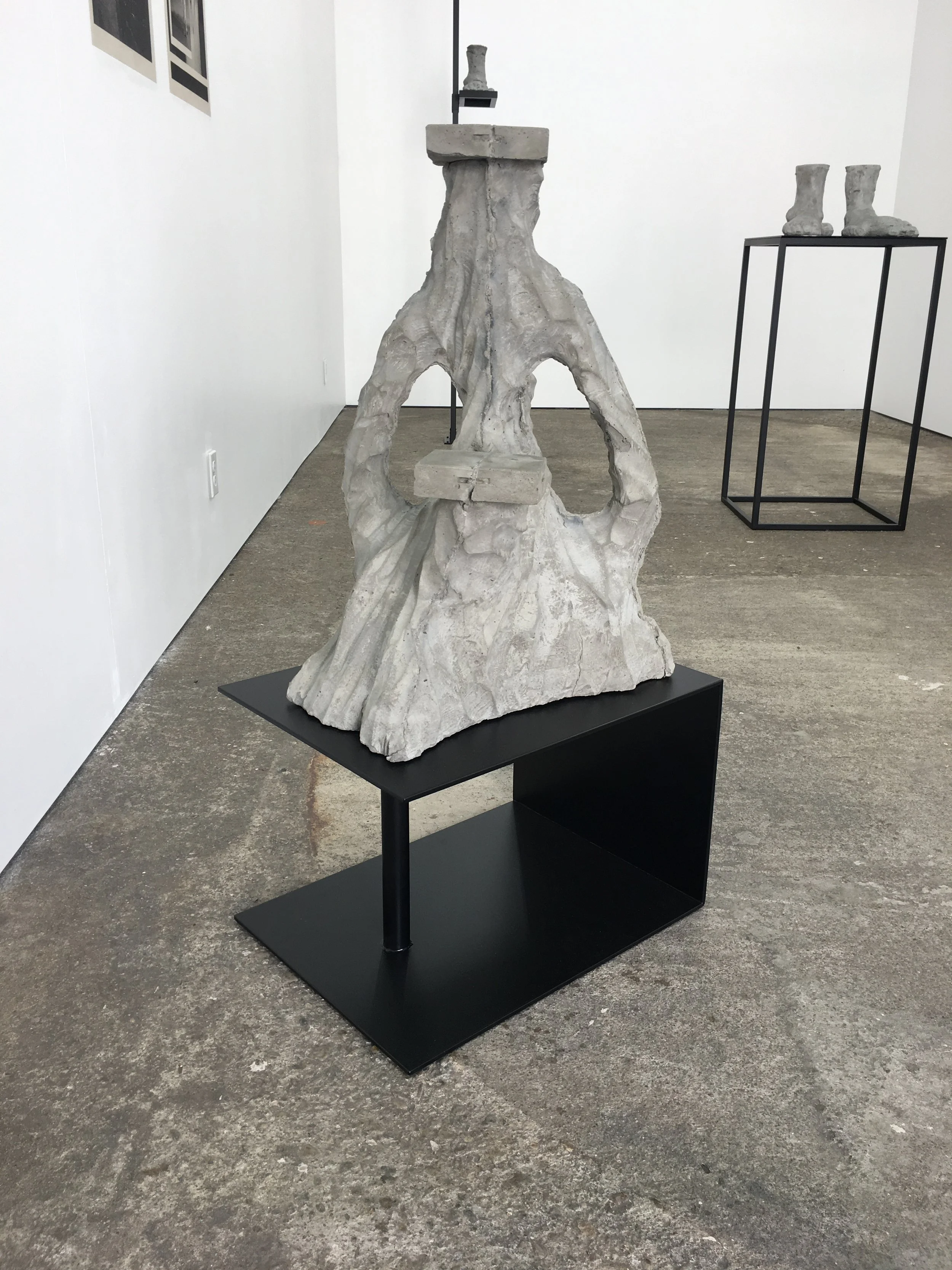 Sculptural artworks in a gallery setting, featuring a textured, abstract sculpture on a black stand in the foreground and another set of sculptures on a metal frame in the background.