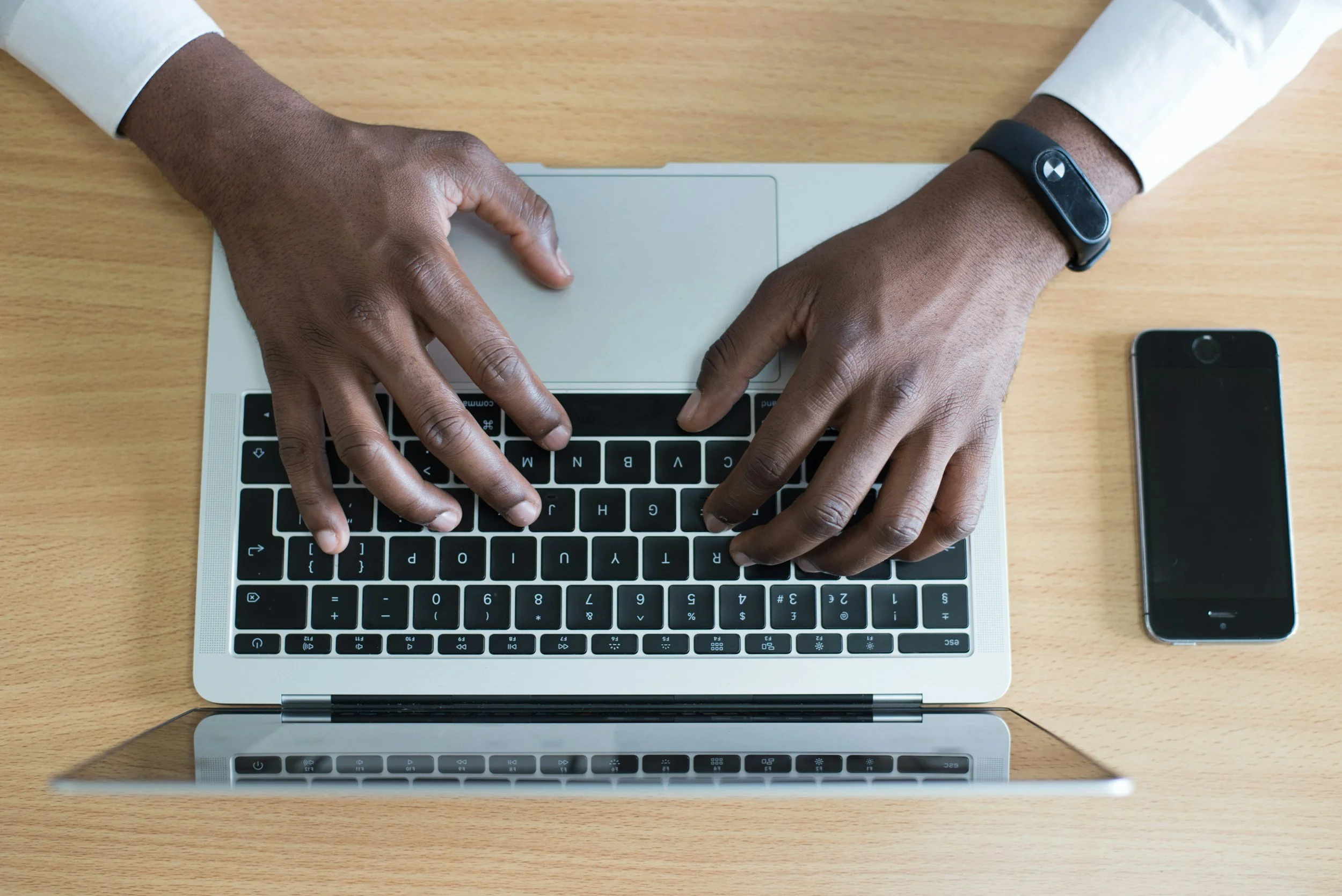 Someone typing on a silver laptop with a black keyboard, a smartphone to the right, and a wooden desk.