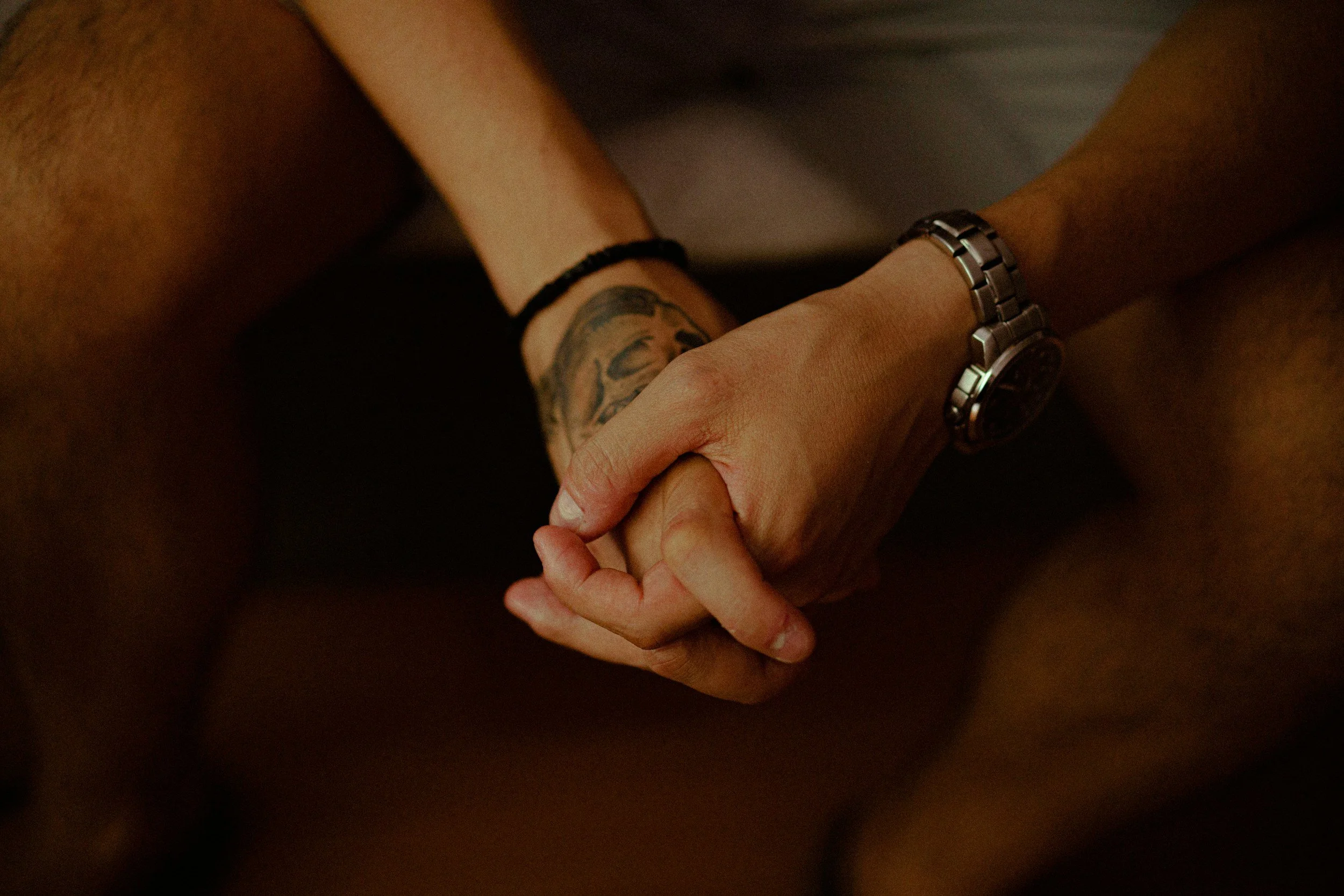 Therapy with Monica / Couples therapy. A close-up of two people holding hands, with one person's arm displaying tattoos and wearing a watch and a black bracelet.