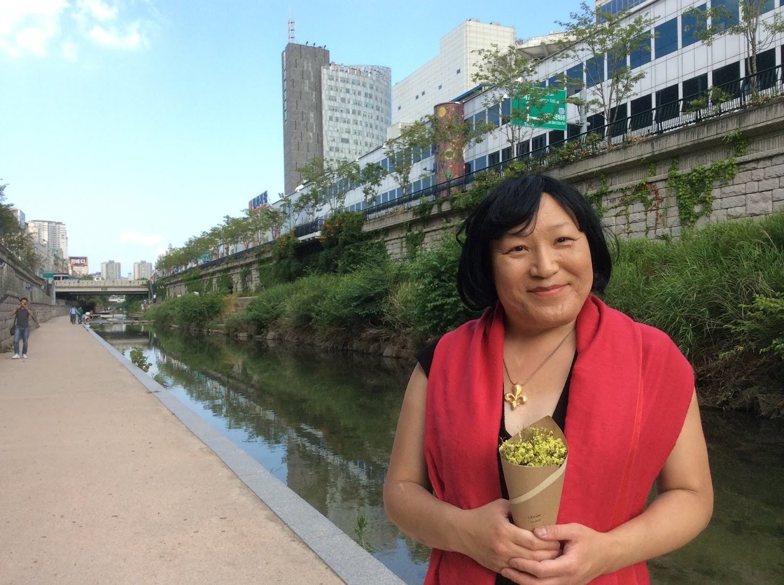 A woman in a black sleeveless top with a red shawl holding a small bouquet of yellow flowers standing near a river in an urban park with tall buildings and trees in the background.