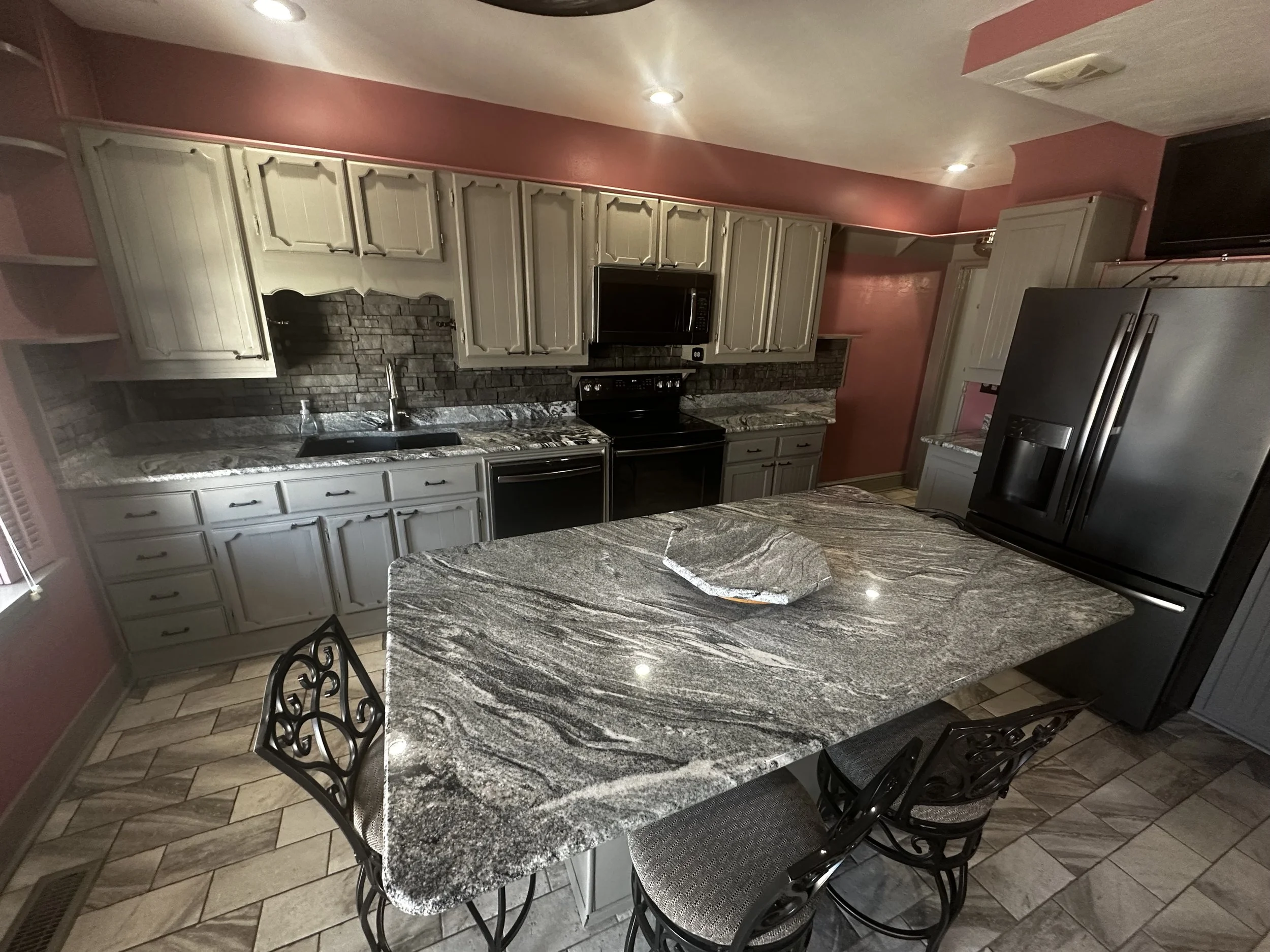 A kitchen with gray cabinets, a granite countertop and island, black appliances, and pink walls.