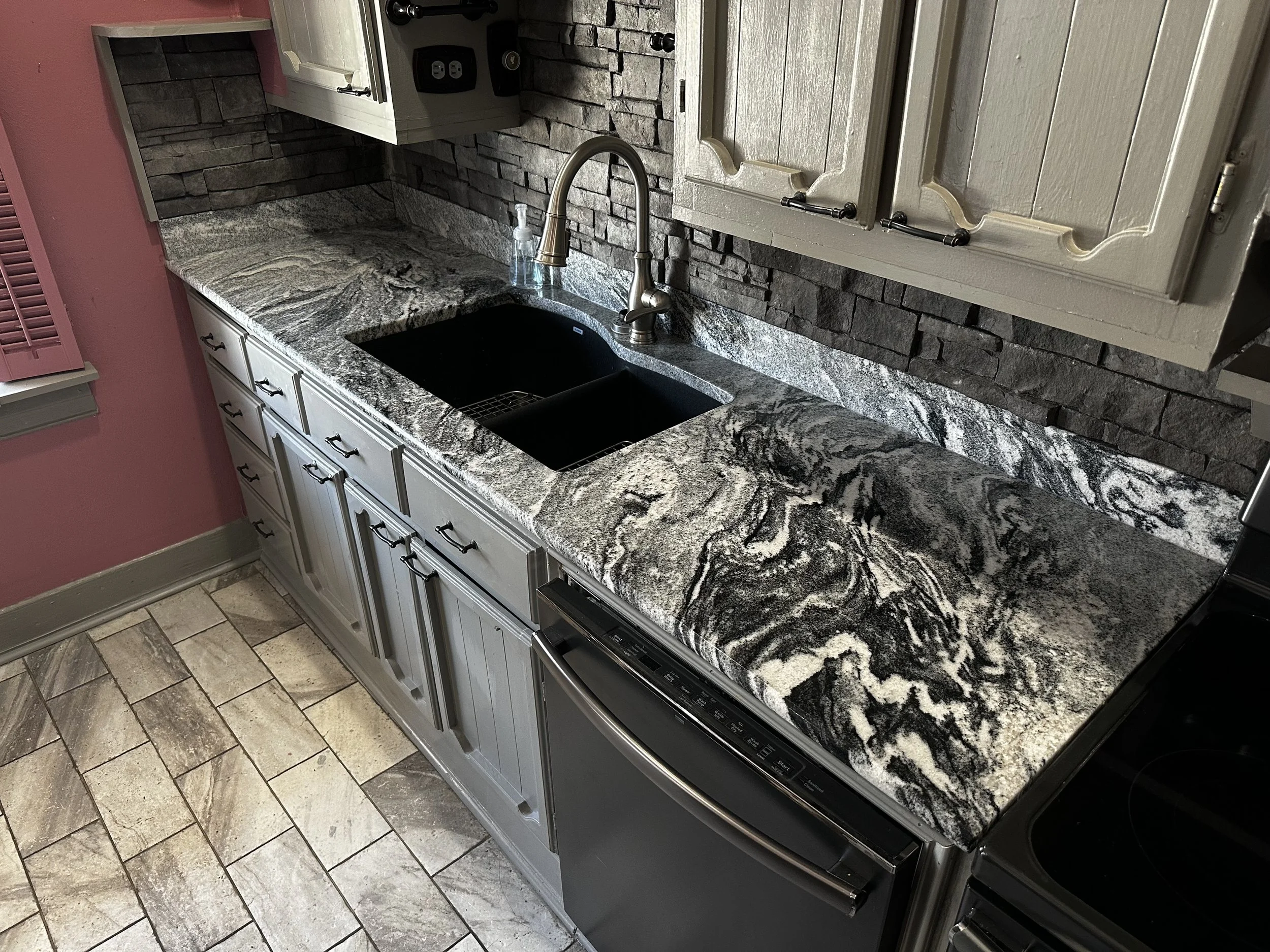 Kitchen countertop with black double sink, granite surface with black, white, and gray swirling patterns, beige cabinets, a stone backsplash, and a black stovetop.