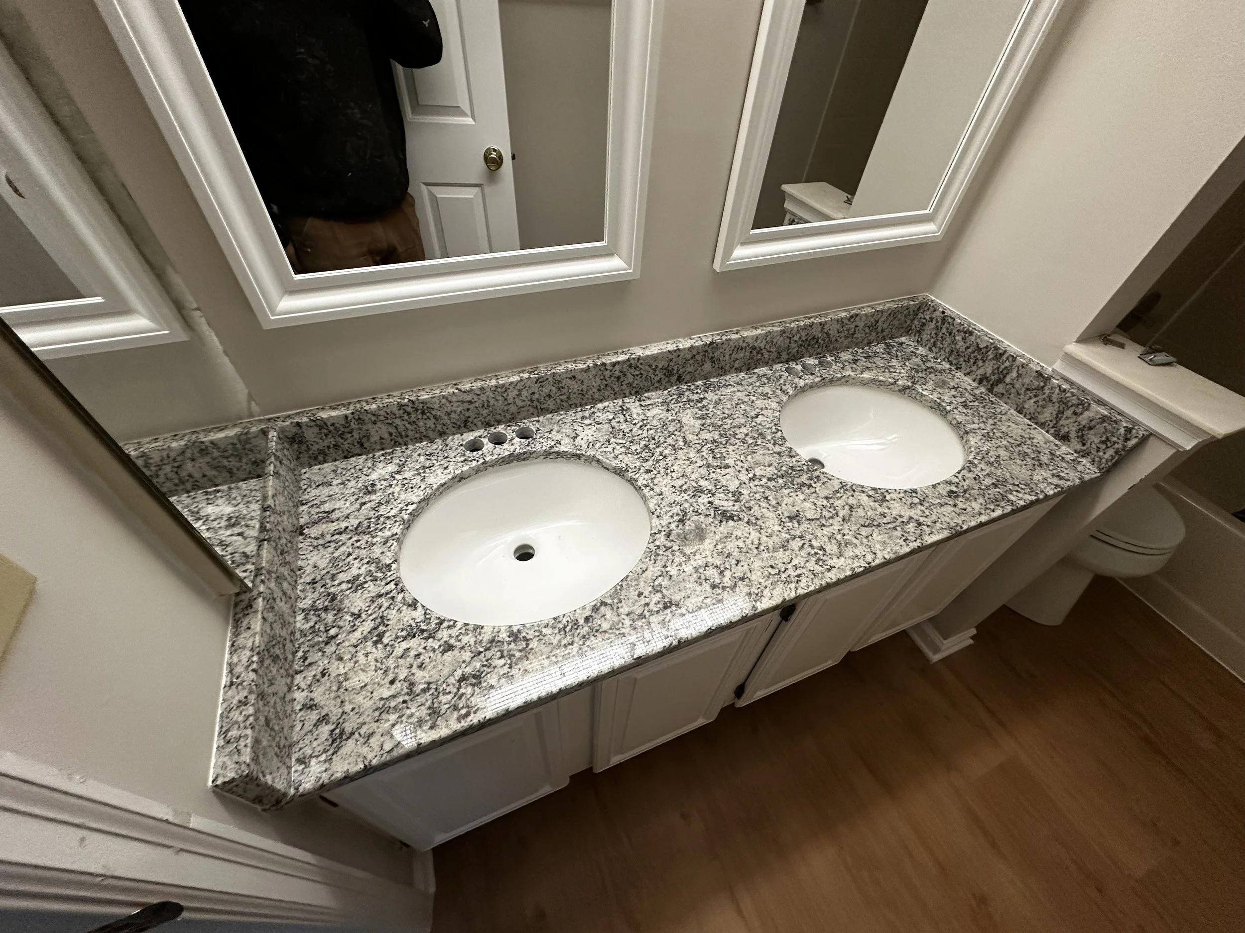 Bathroom vanity with a granite countertop with two oval undermount sinks, positioned beneath two mirrors, and visible reflection of a door and a person in a black shirt.