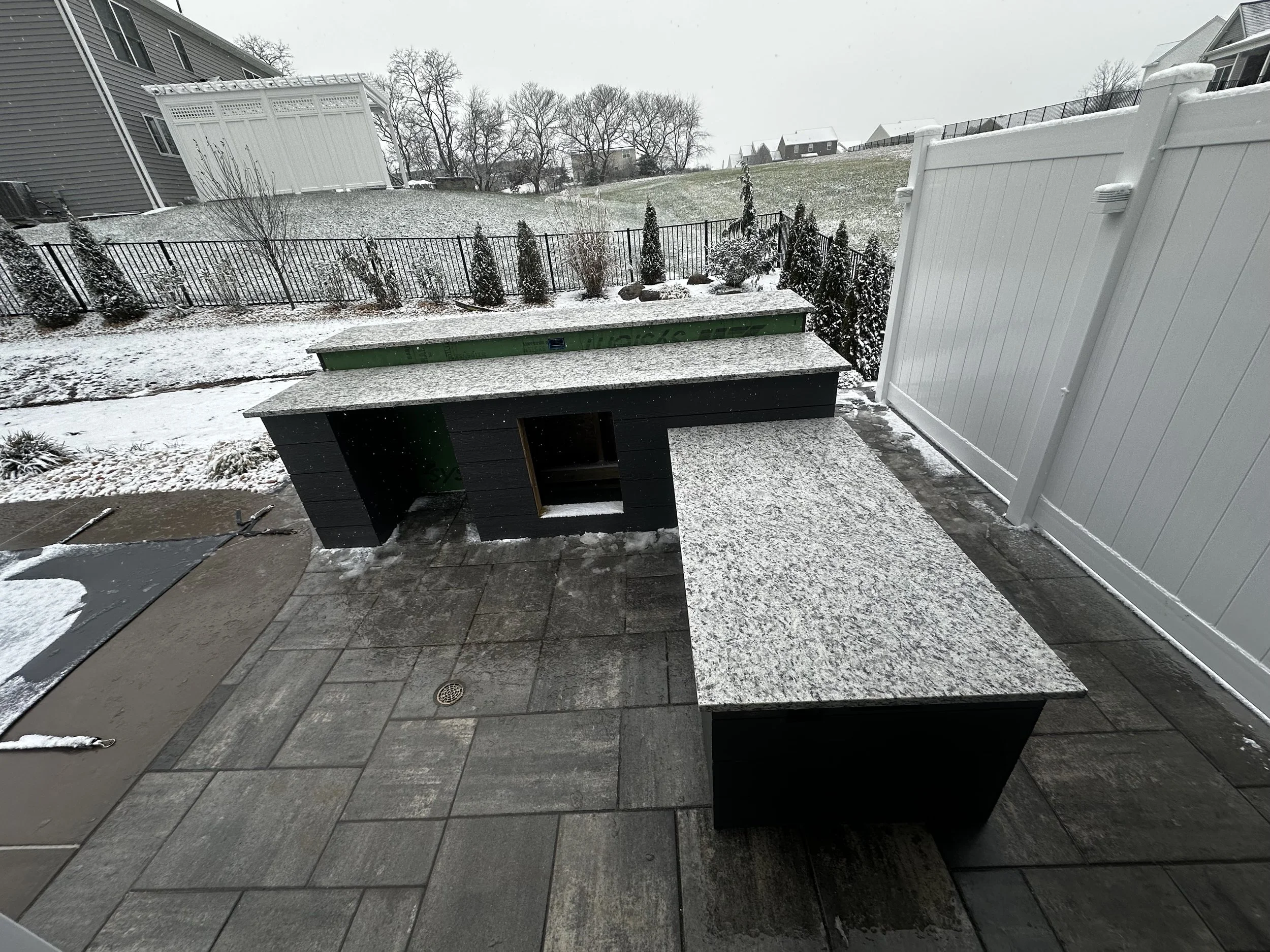 Outdoor patio with black and granite countertops, snow-covered ground, fenced backyard, and snow falling in a residential neighborhood.