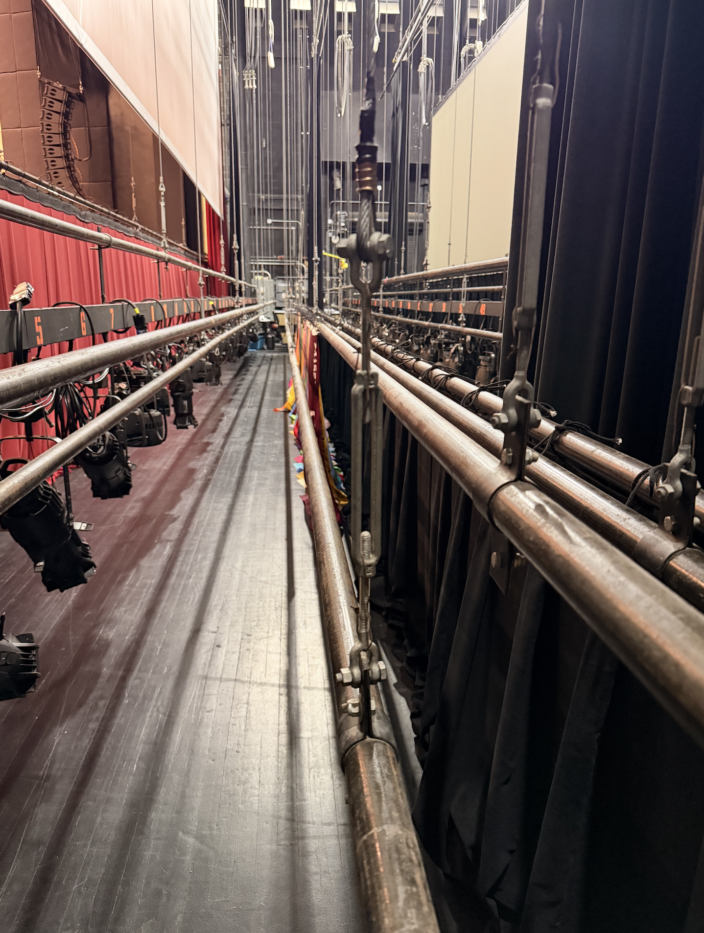 View from behind stage lighting and rigging equipment, with red stage curtain visible on the left and black curtains on the right side of the image.