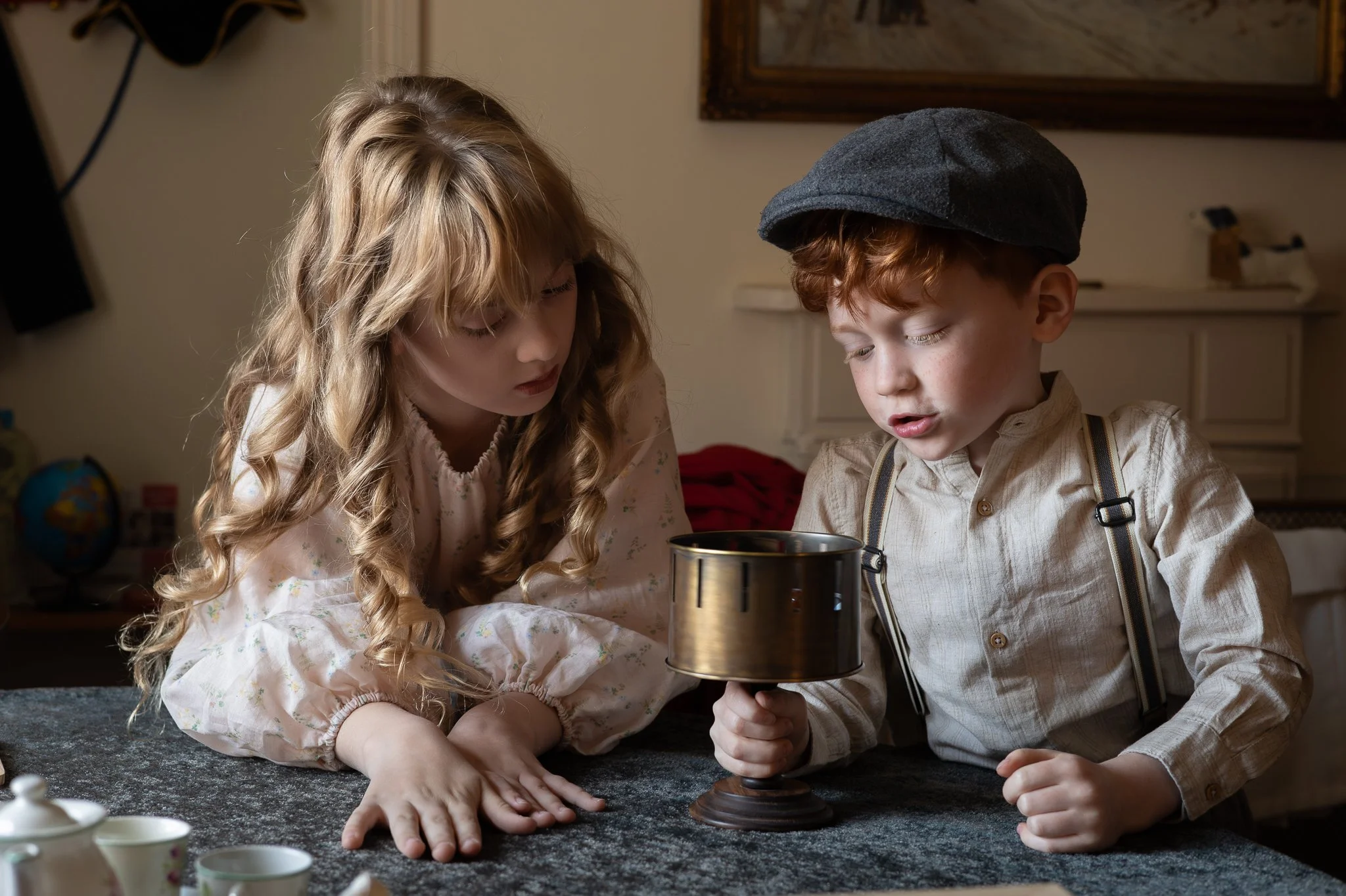 A young girl and boy with light skin sitting on a table, looking at a vintage telescope. The girl has long, curly blonde hair and is wearing a light-colored dress with floral patterns. The boy has short, curly red hair, is wearing a beige shirt, suspenders, and a gray flat cap. The background shows a beige wall with a framed picture, a white dresser, and some objects including a globe and cups.