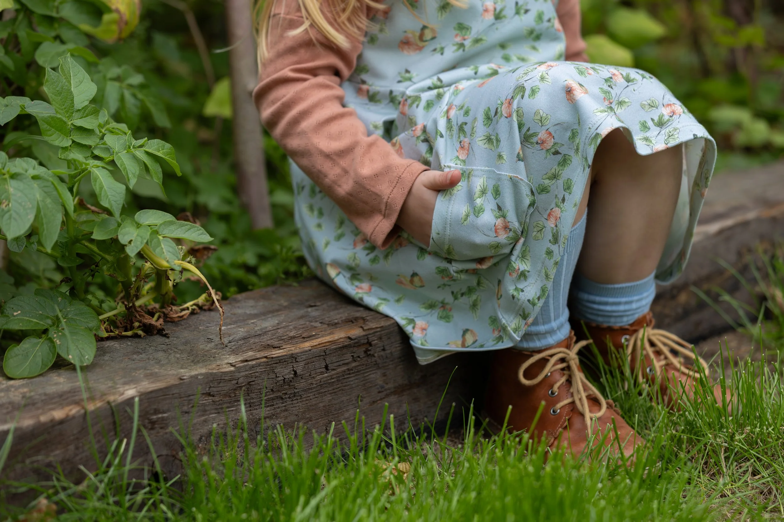 Child in floral dress sitting on a tree log outdoors with green grass and plants nearby.
