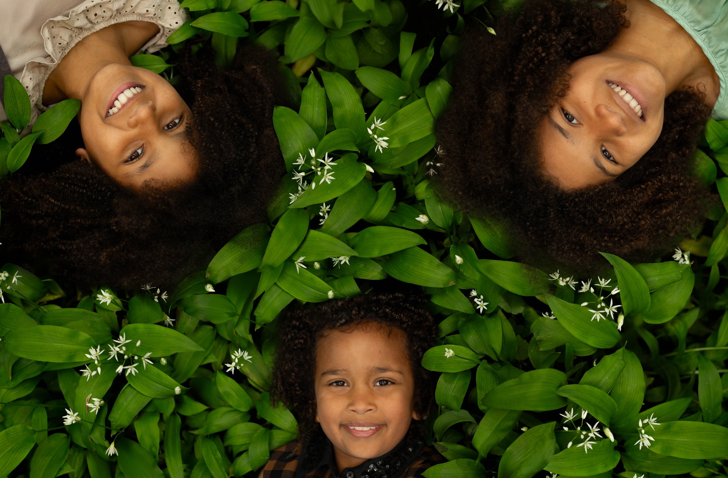 Three people lying on green leaves with small white flowers, smiling, heads towards each other.