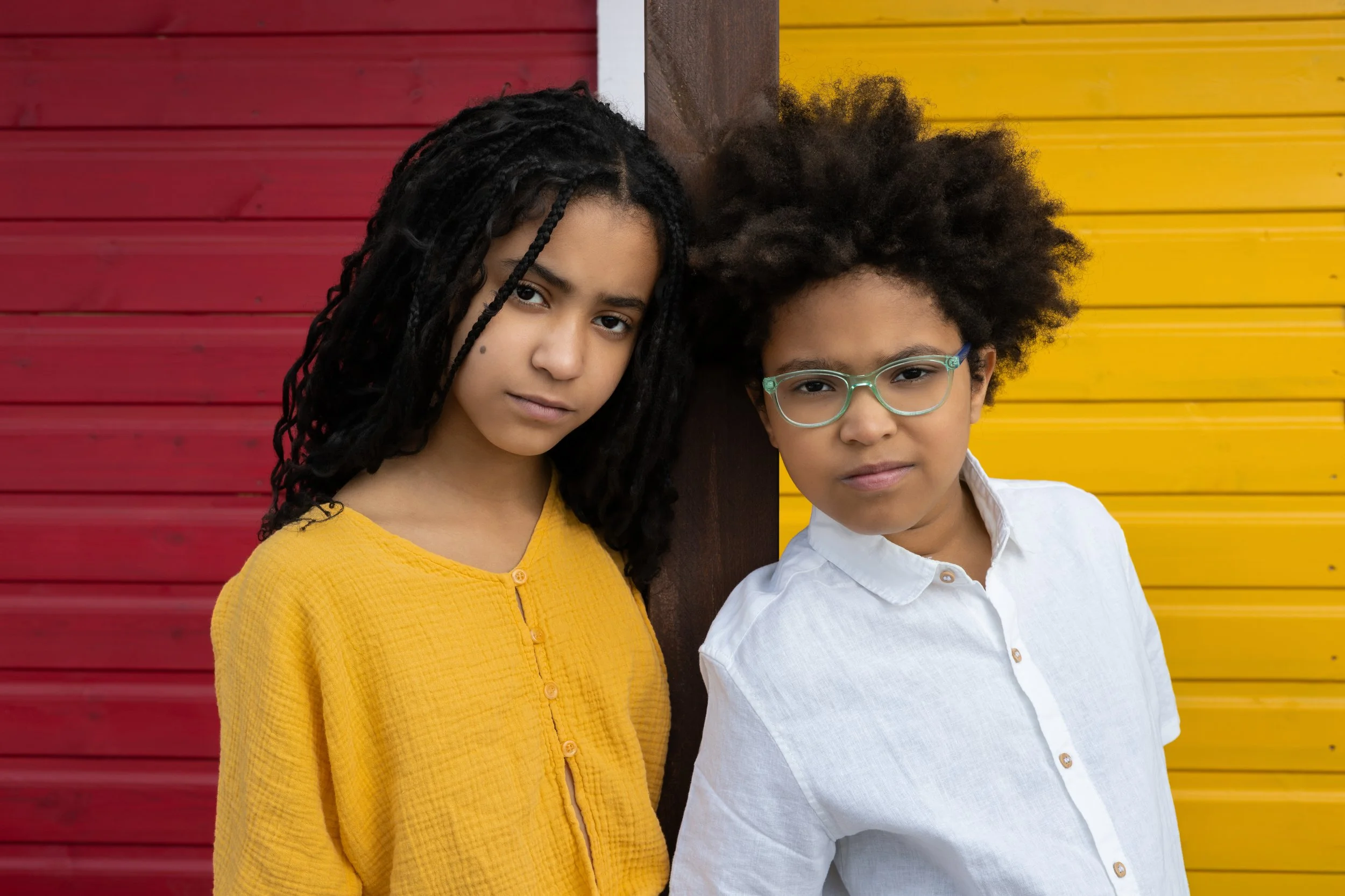Two children, a girl and a boy, stand close together against a colorful background with red, brown, and yellow wooden panels. The girl, on the left, has long braided hair and wears a mustard yellow top, with a serious expression. The boy, on the right, has curly hair, wears light blue glasses and a white shirt, and has a neutral expression.