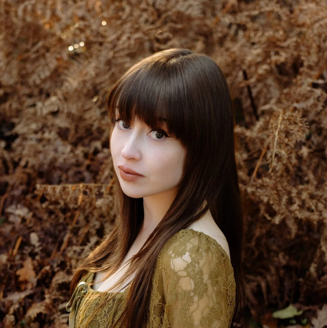 A young woman with long brown hair and bangs posing outdoors with a background of dry brown leaves.