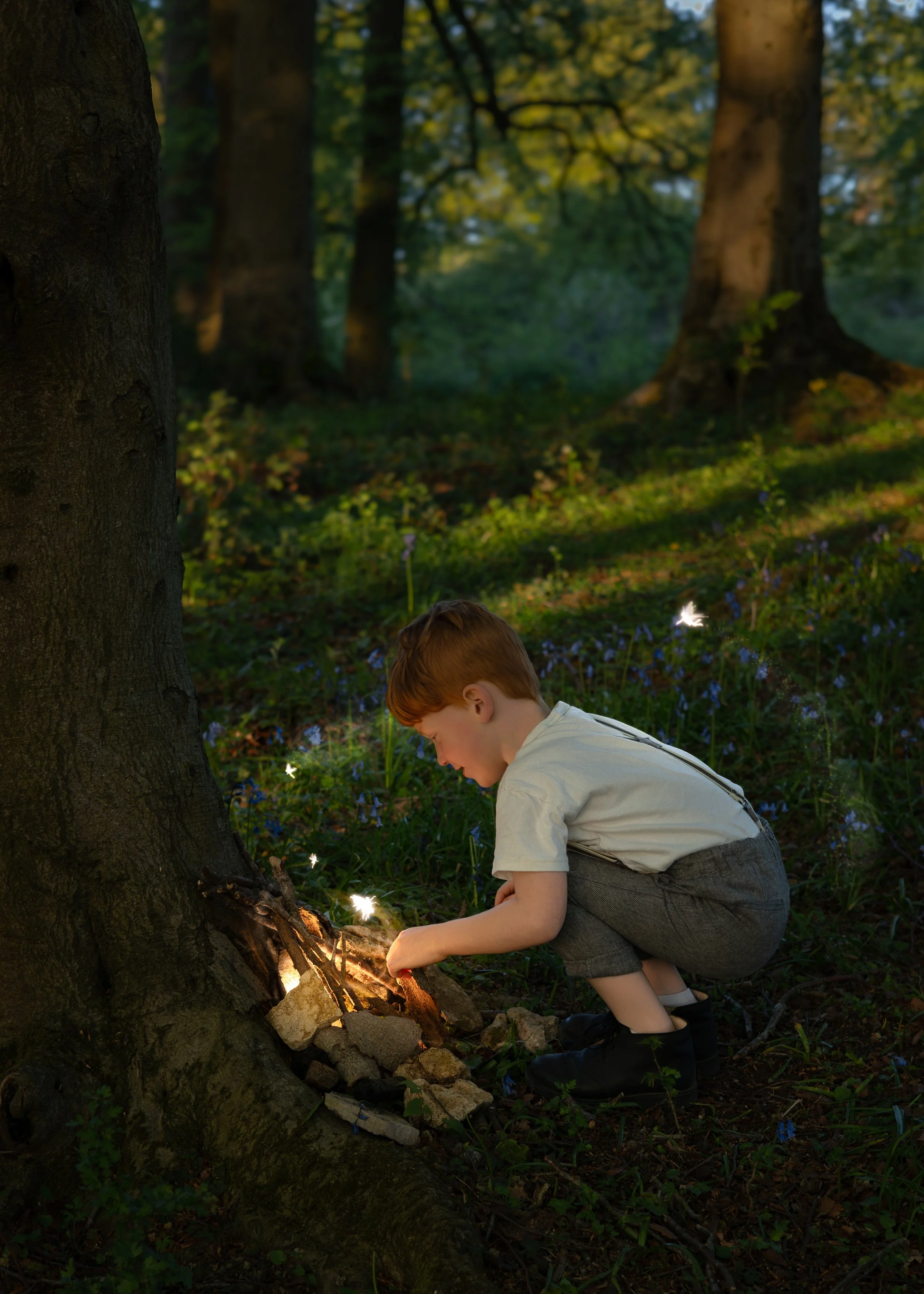 A young boy with red hair, wearing a white shirt and gray shorts, is crouching by a tree in a forest during daytime. He is near a small campfire with rocks, and sparks are visible, with green foliage and small blue flowers surrounding him.