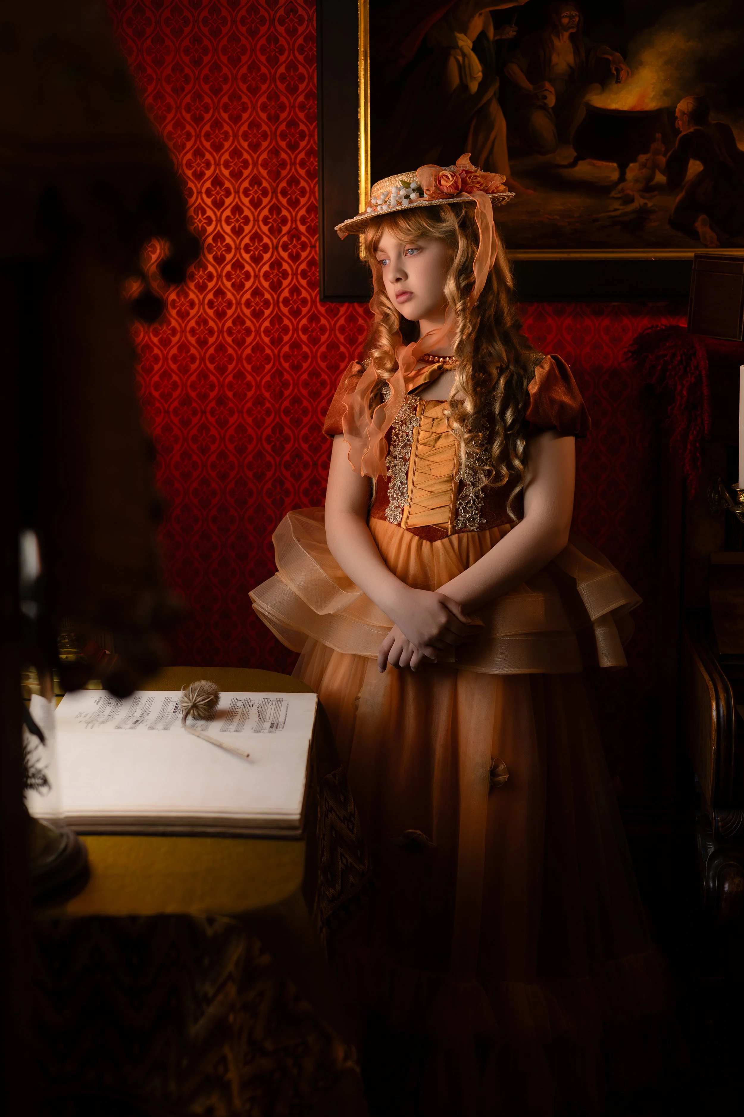 A young girl dressed in vintage, Victorian-style clothing, with a ribboned hat, stands solemnly inside a dimly lit room with red wallpaper and a dark painting in the background. She appears to be in a serious or contemplative mood, with her hands clasped in front of her.