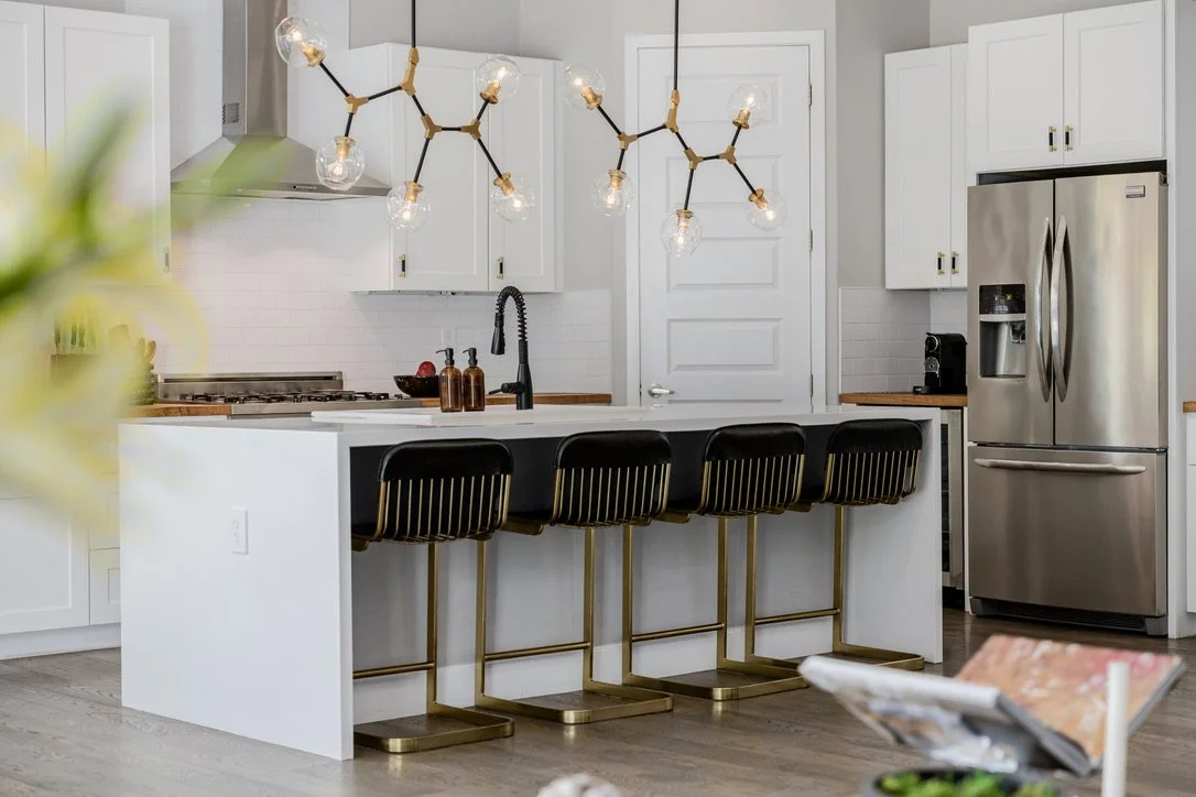 Modern kitchen with white island, four black bar stools, brass lighting fixtures, and stainless steel appliances.