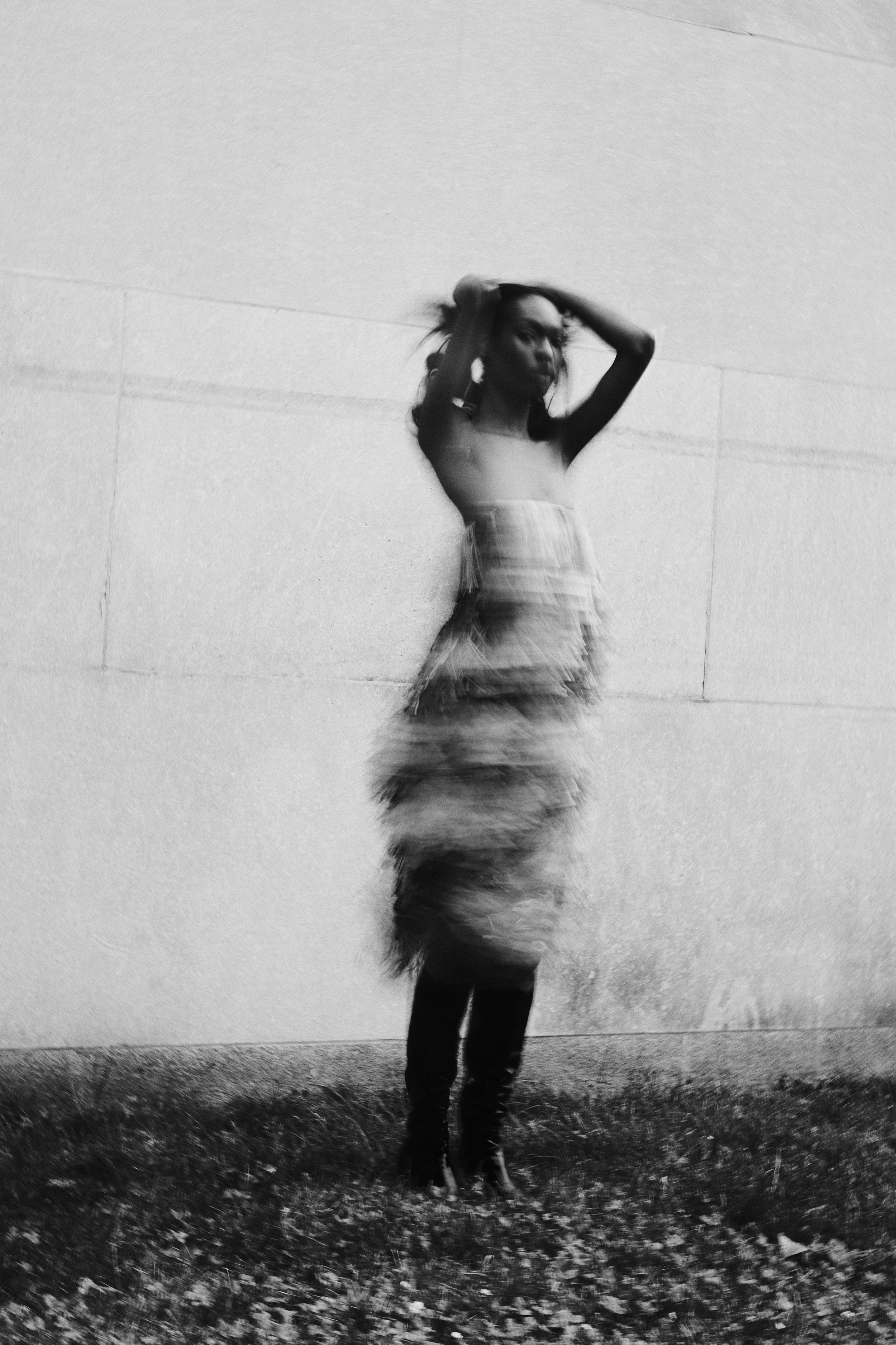 Blurred black and white image of a person in motion with arms raised, wearing a dress and standing on grass.