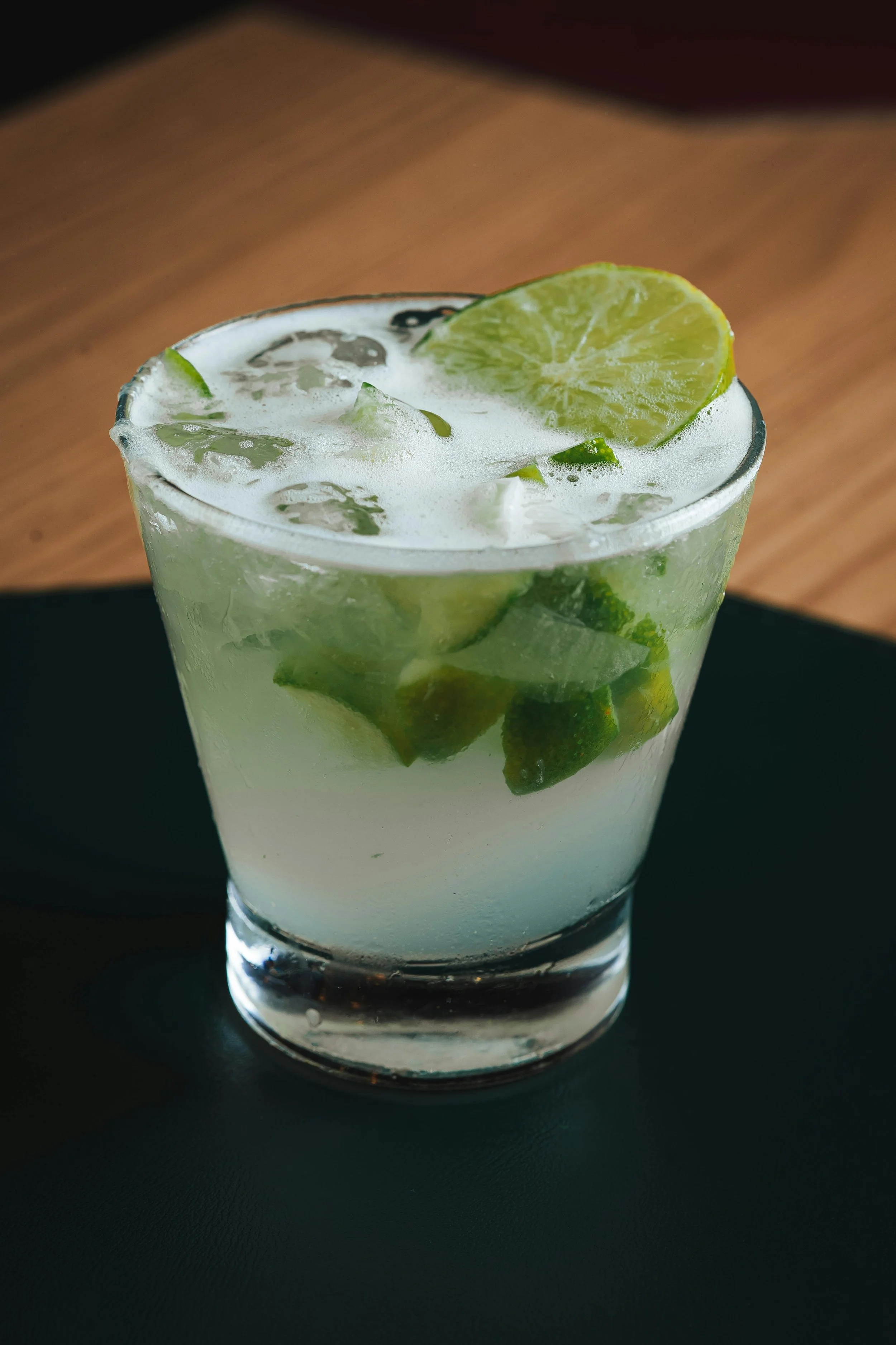 A close-up image of a glass of limeade with ice and a lime wedge garnish on top, sitting on a black surface with a wooden background.