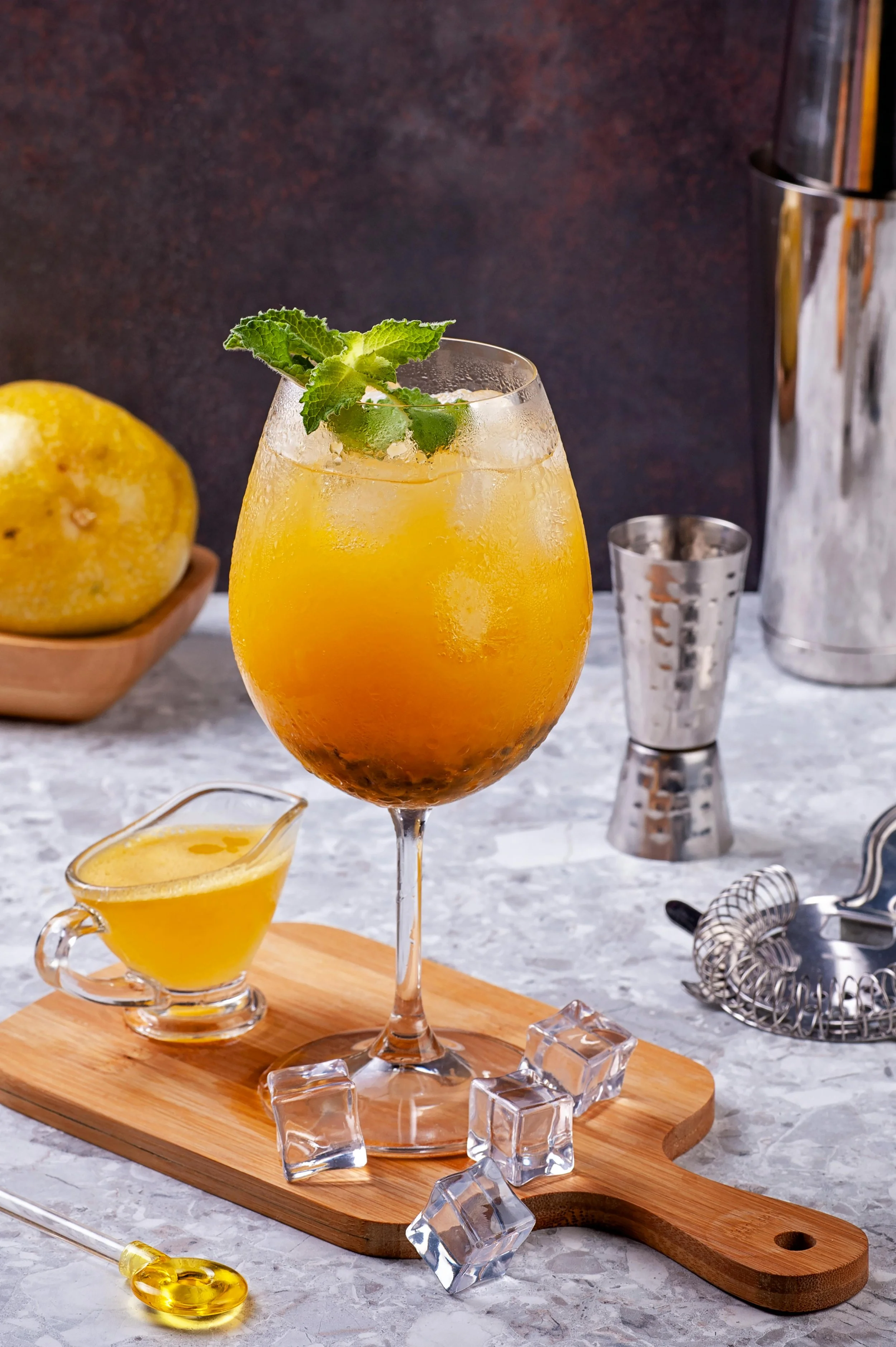 A cocktail with ice, garnished with mint, on a wooden board surrounded by ice cubes and a small pitcher of lemon juice, with a lemon in the background.