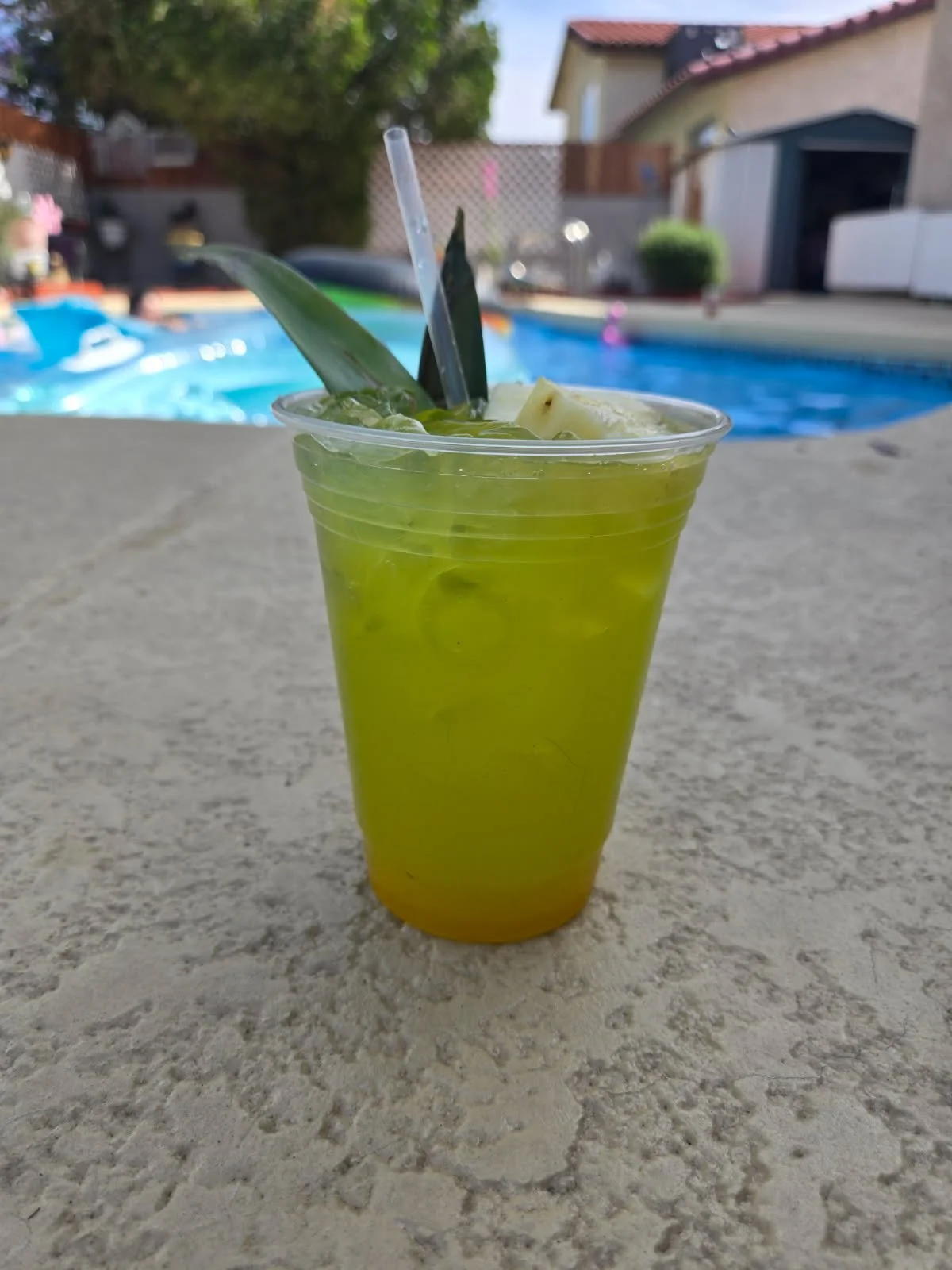A tropical drink with pineapple leaves and pineapple chunks in a clear plastic cup on a stone surface near a swimming pool.
