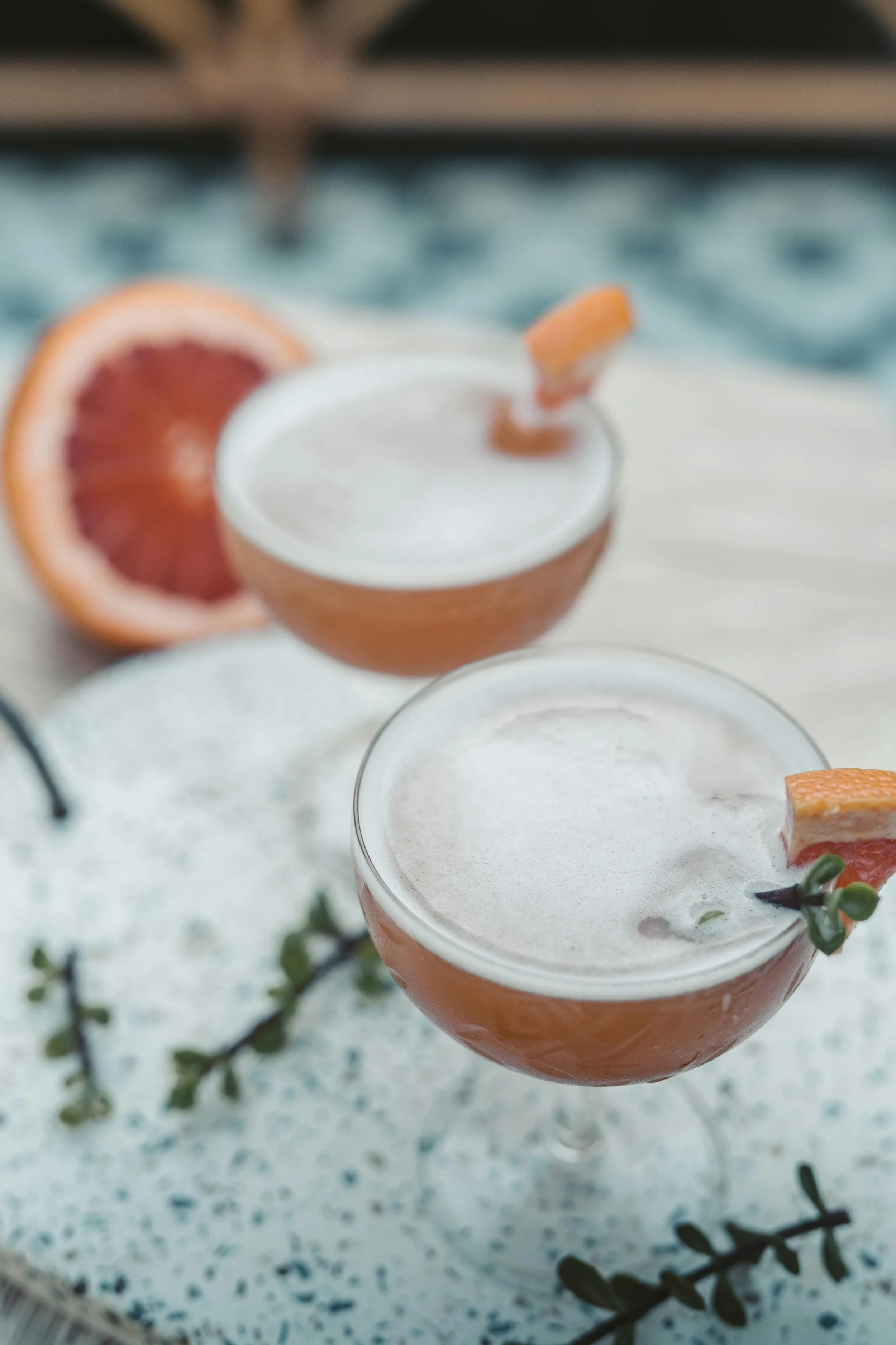 Two grapefruit cocktails garnished with grapefruit wedges and herbs on a white speckled tray with a whole grapefruit in the background and a blue patterned cloth.
