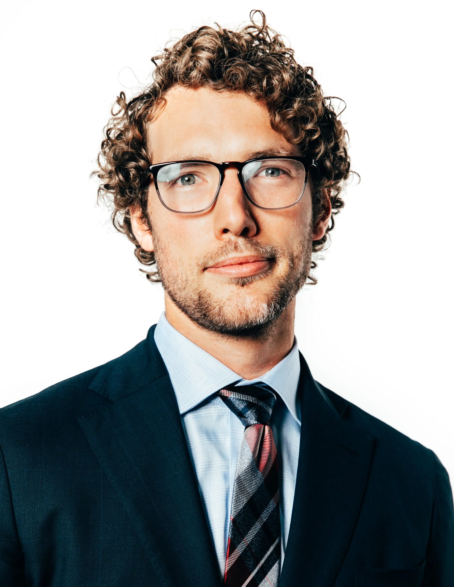 A man with curly hair, glasses, wearing a suit and tie, posing against a white background.