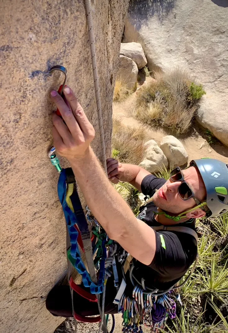 A man rock climbing on a steep rock face, wearing a helmet, sunglasses, and climbing gear, reaching for a handhold with one hand while holding a safety rope with the other.