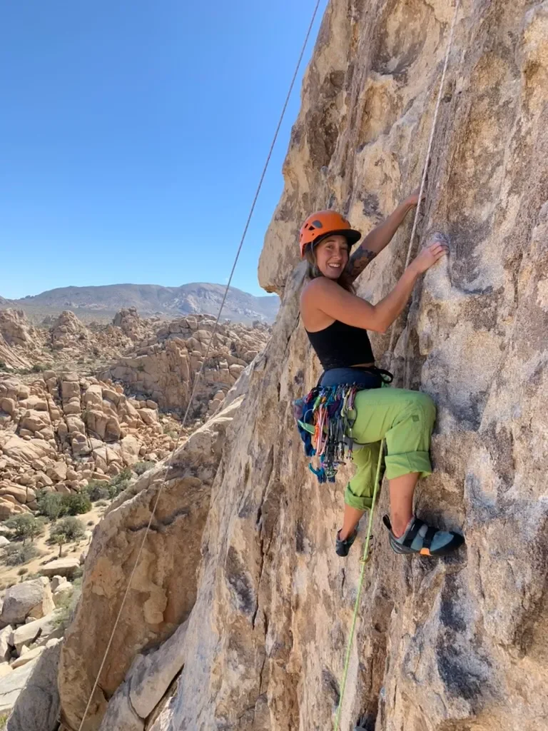 A woman is rock climbing on an outdoor cliff, wearing a helmet, climbing shoes, and climbing gear, smiling at the camera.
