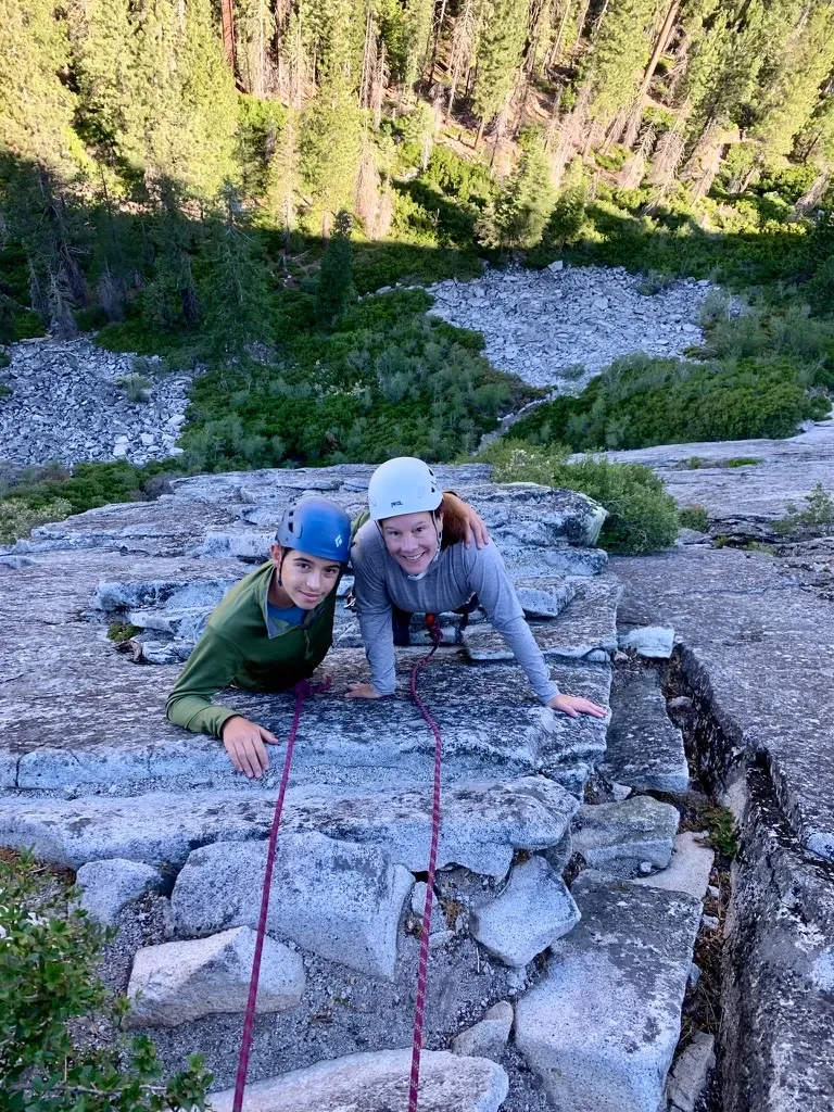Two people in helmets rock climbing on a steep granite surface with a forested mountain landscape in the background.