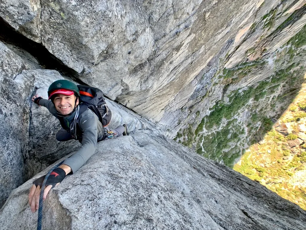 A man rock climbing on a steep granite cliff, wearing a helmet and harness, hanging upside down.