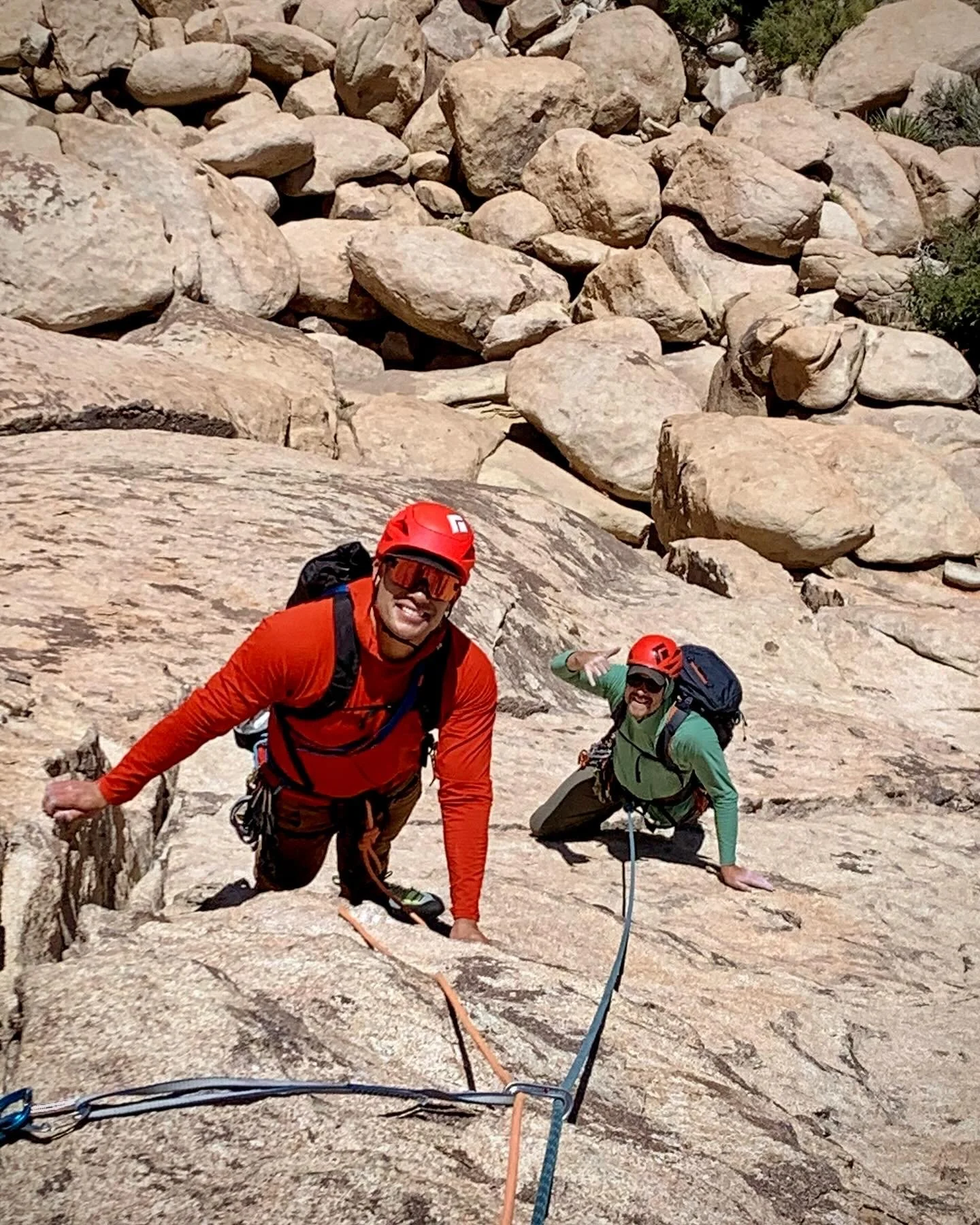 Two climbers wearing helmets and harnesses on a steep rock face, with one climber smiling at the camera and the other making a peace sign, surrounded by large boulders.