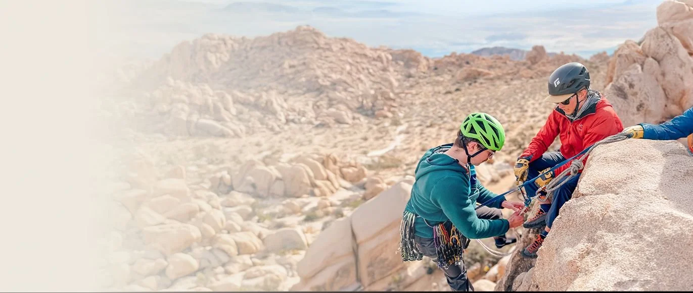 Two people helping a child climb a rock face in a desert landscape, all wearing helmets and safety gear.
