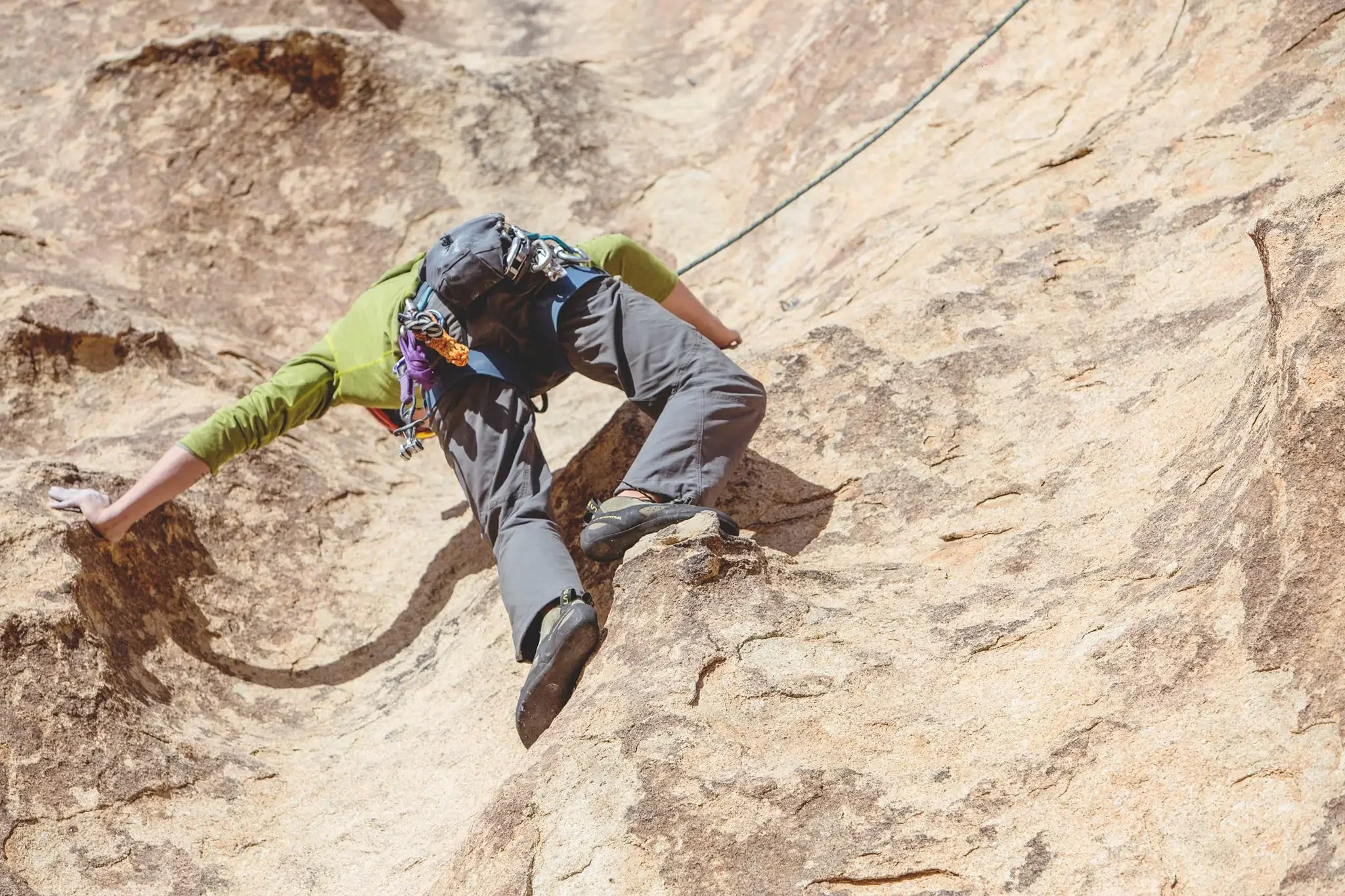 A person rock climbing on a steep, rocky cliff. They are wearing a green shirt, gray pants, and climbing shoes, with a harness and safety gear, ascending upward.