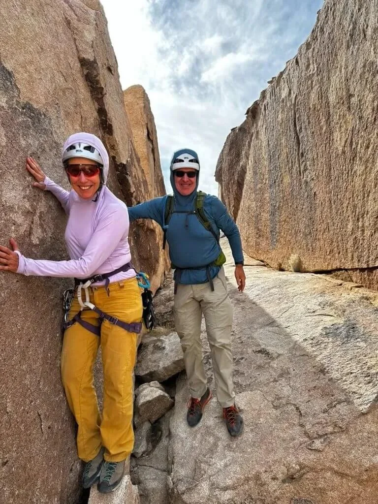 Two women in outdoor climbing gear, helmets, and sunglasses smiling while climbing between large rock formations.