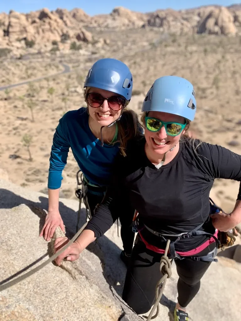 Two women wearing climbing helmets and sunglasses smiling on a rocky mountain ledge with desert landscape in the background.