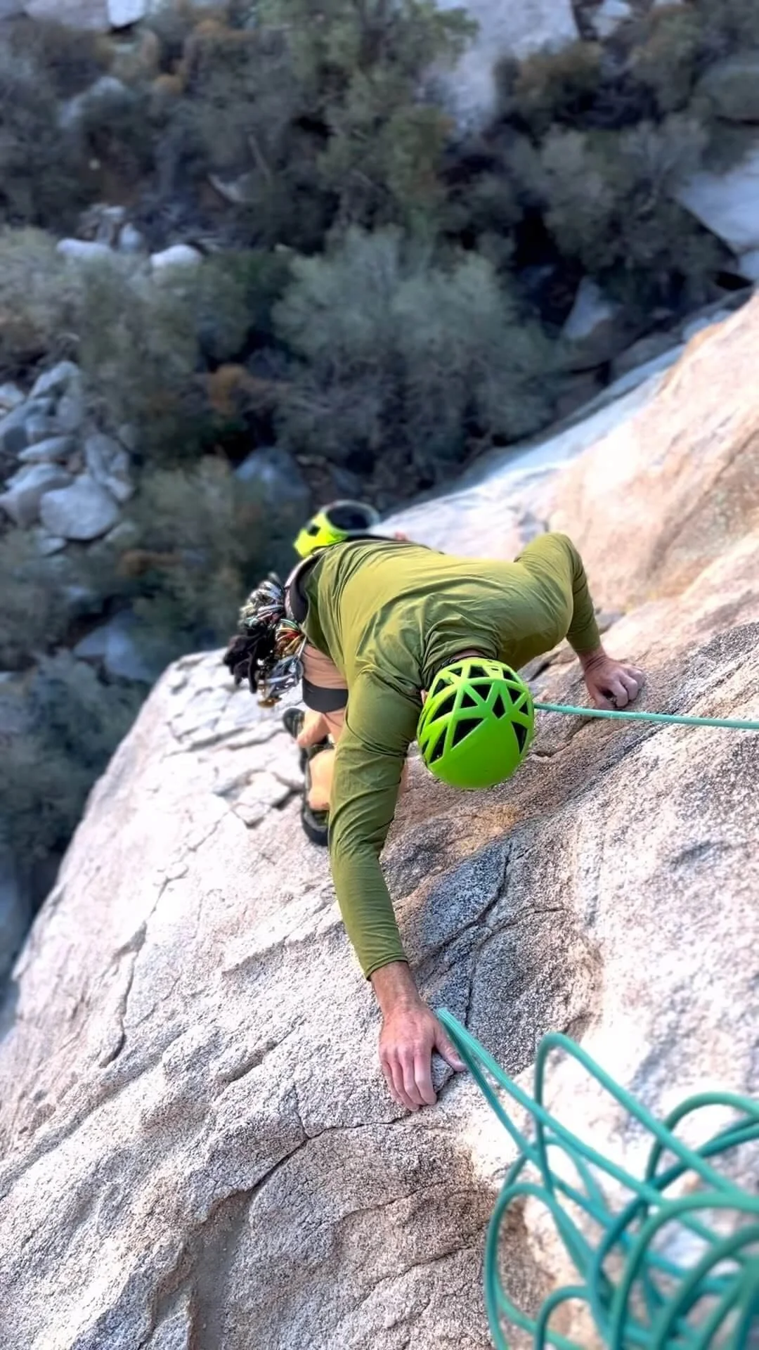 Person climbing a rock face with a green helmet on, using their hands and feet, secured with a safety harness and rope, in an outdoor natural setting.