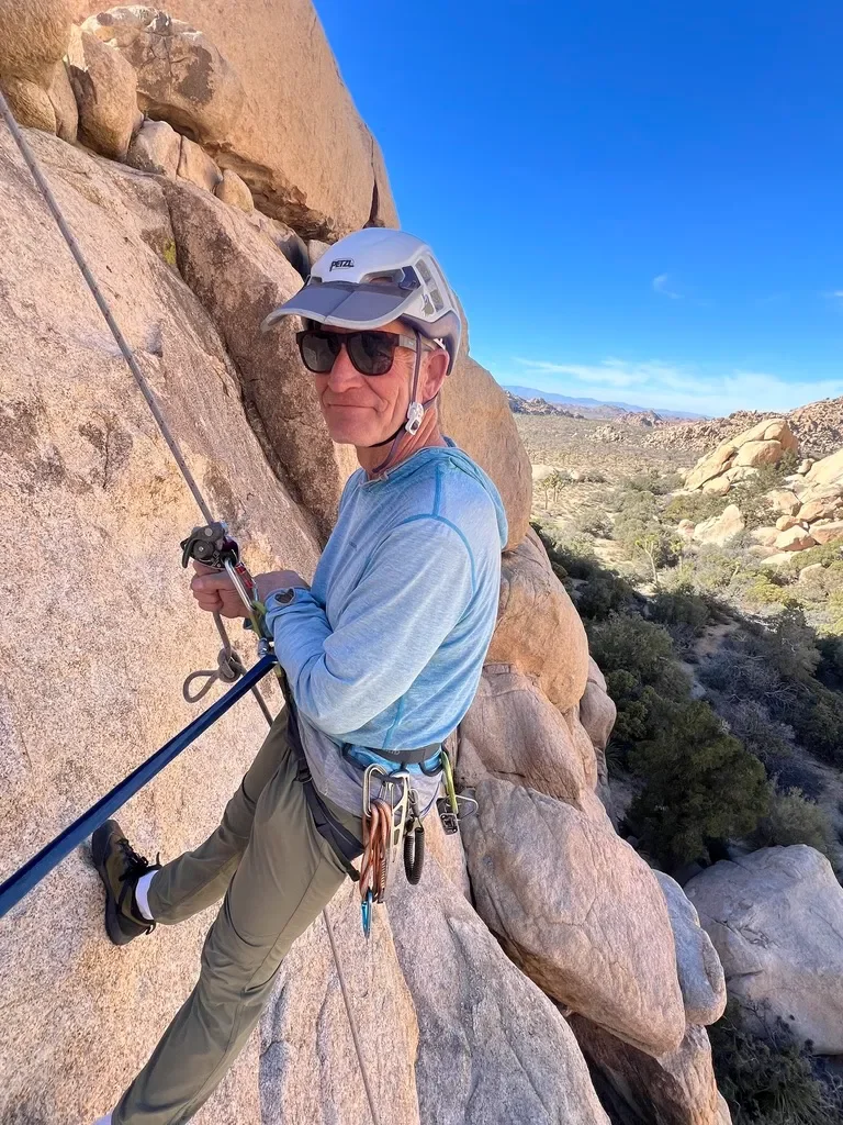 A man rock climbing on a vertical rock face in a desert landscape, wearing a helmet, sunglasses, a long sleeve shirt, climbing harness, and holding a rope.