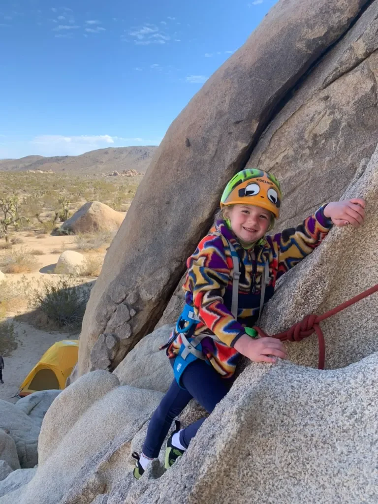 A young girl rock climbing outdoors on a large rock formation, wearing a colorful jacket, a helmet, and a harness, smiling at the camera.