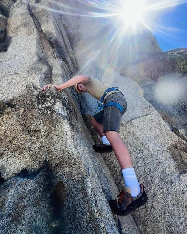A person climbing a steep granite rock face outdoors on a sunny day with a bright sun and clear sky.