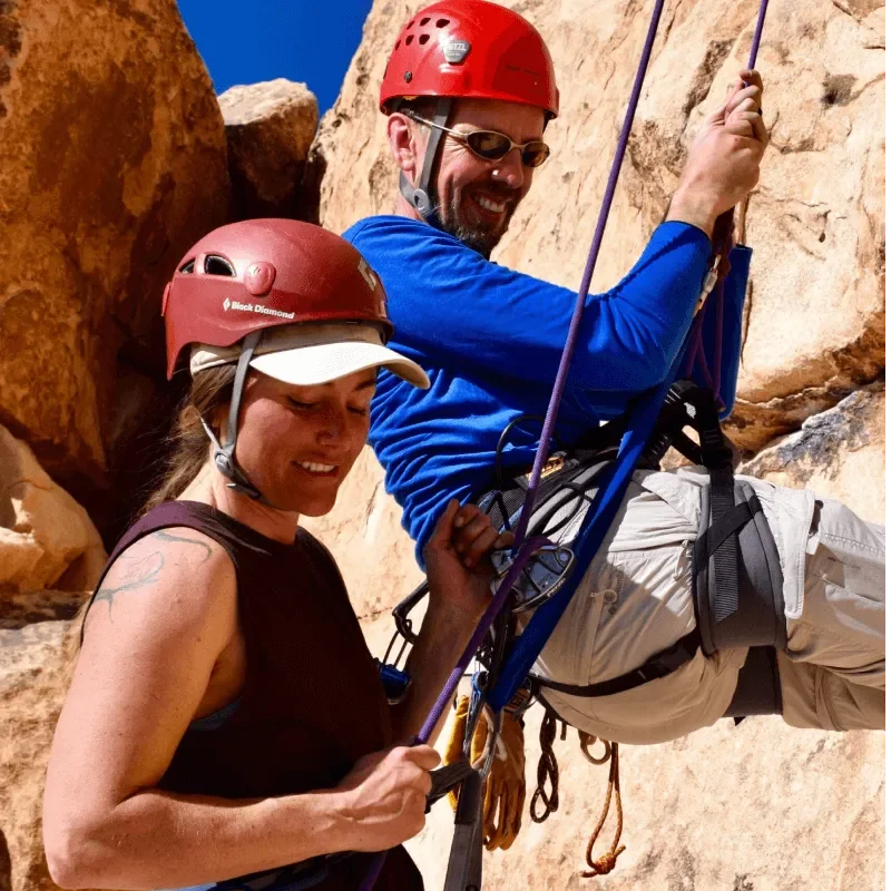A man and woman rock climbing on a rugged outdoor cliff face, both wearing helmets and harnesses.